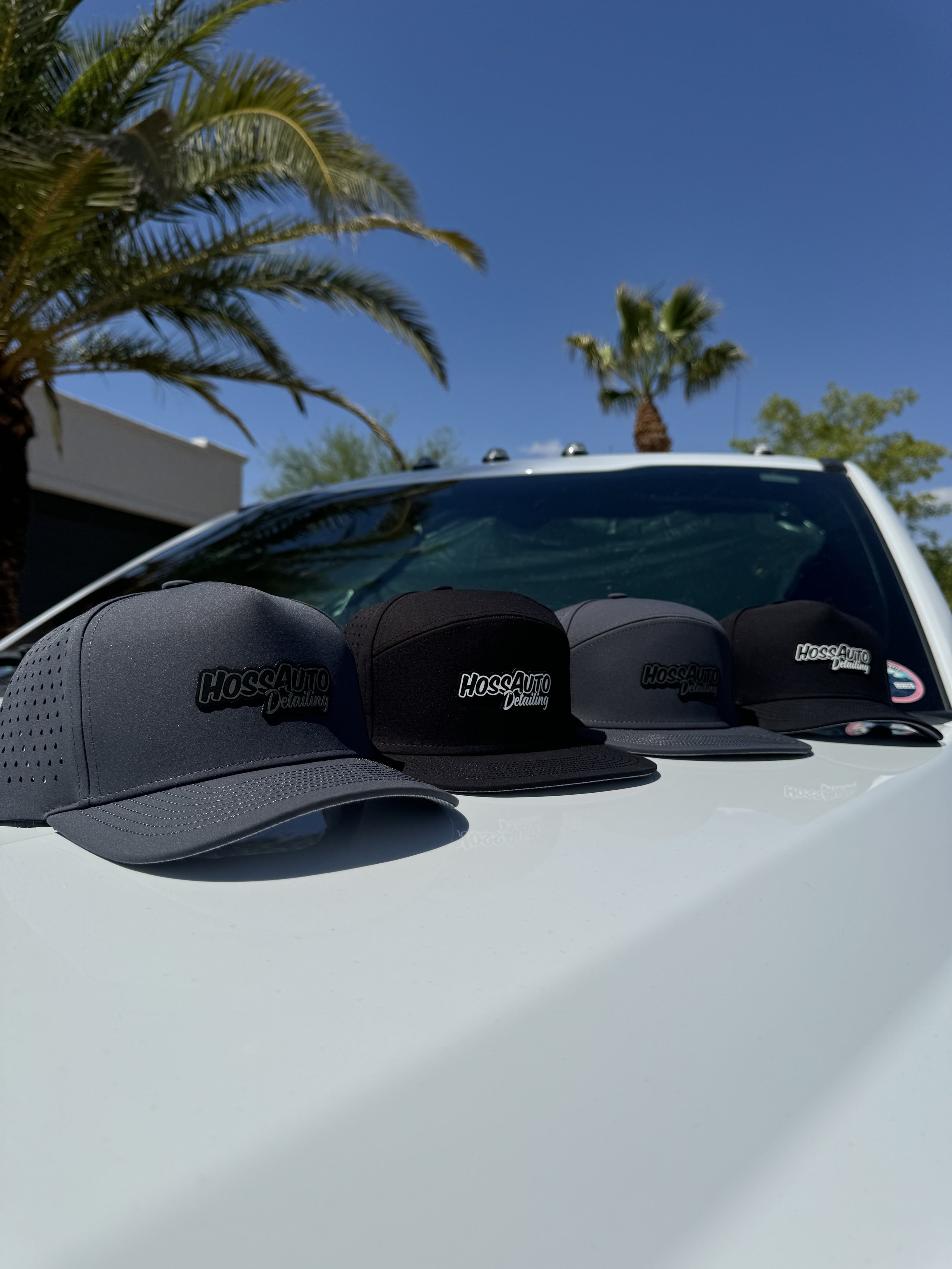Four black baseball caps with the logo 'HossAuto Detailing' on a white vehicle hood, with palm trees and a clear blue sky in the background.