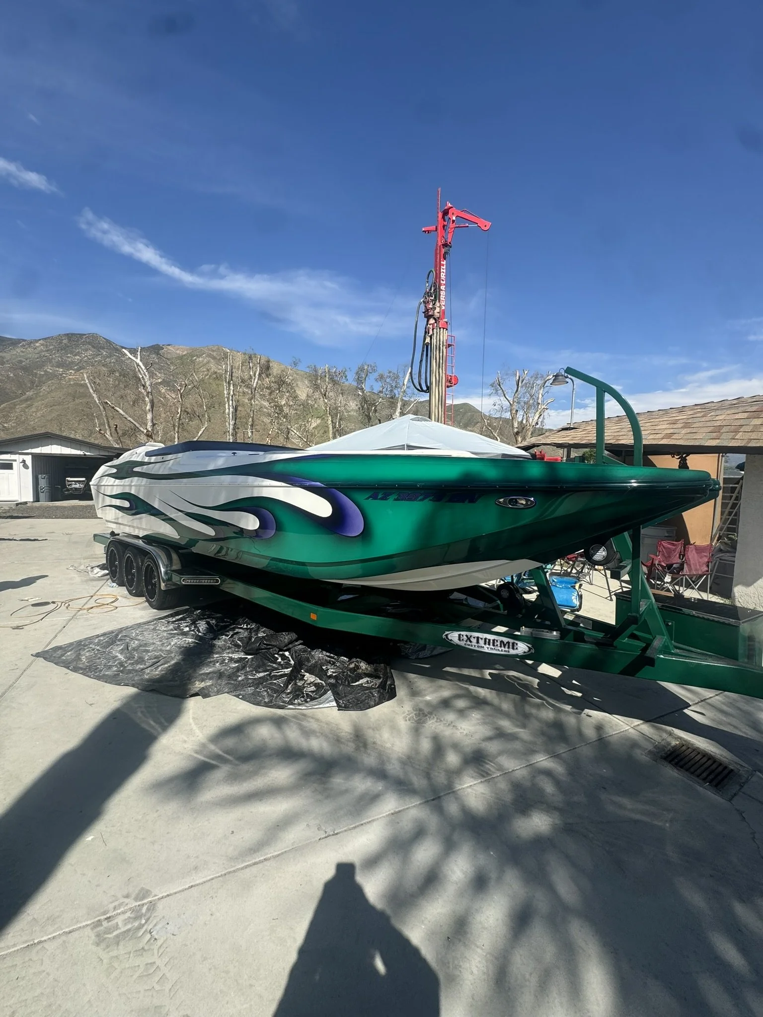 A speedboat on a green trailer parked outside on concrete, with a mountainous background and a blue sky. The boat has a sleek design with black and purple flame graphics.