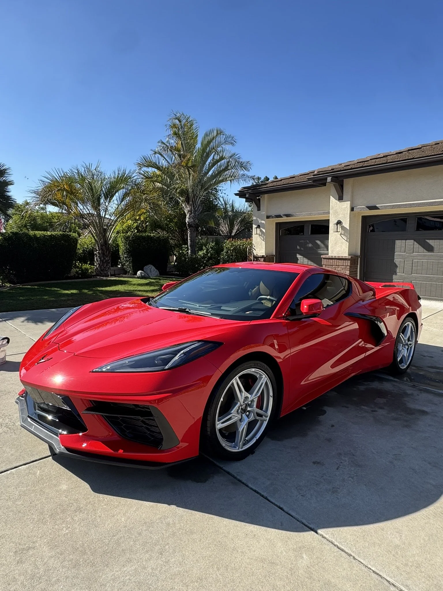 A red sports car parked in front of a house with a beige exterior, brown roof, garage doors, and a landscaped yard with palm trees.