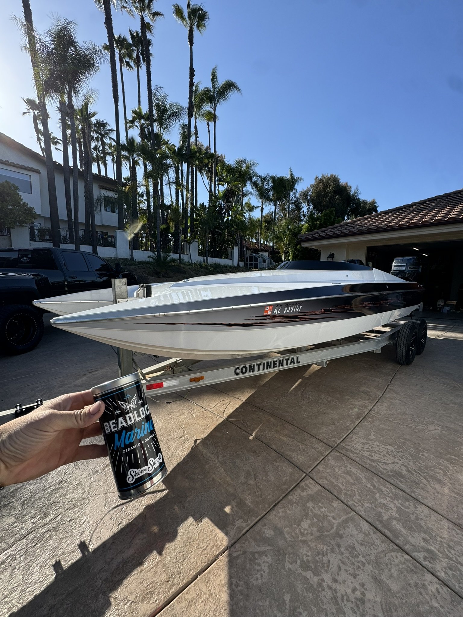 A person holds a can of Beadlock Marine ceramic coating in front of a trailer with a white boat, parked in a driveway with palm trees and houses in the background during daytime.