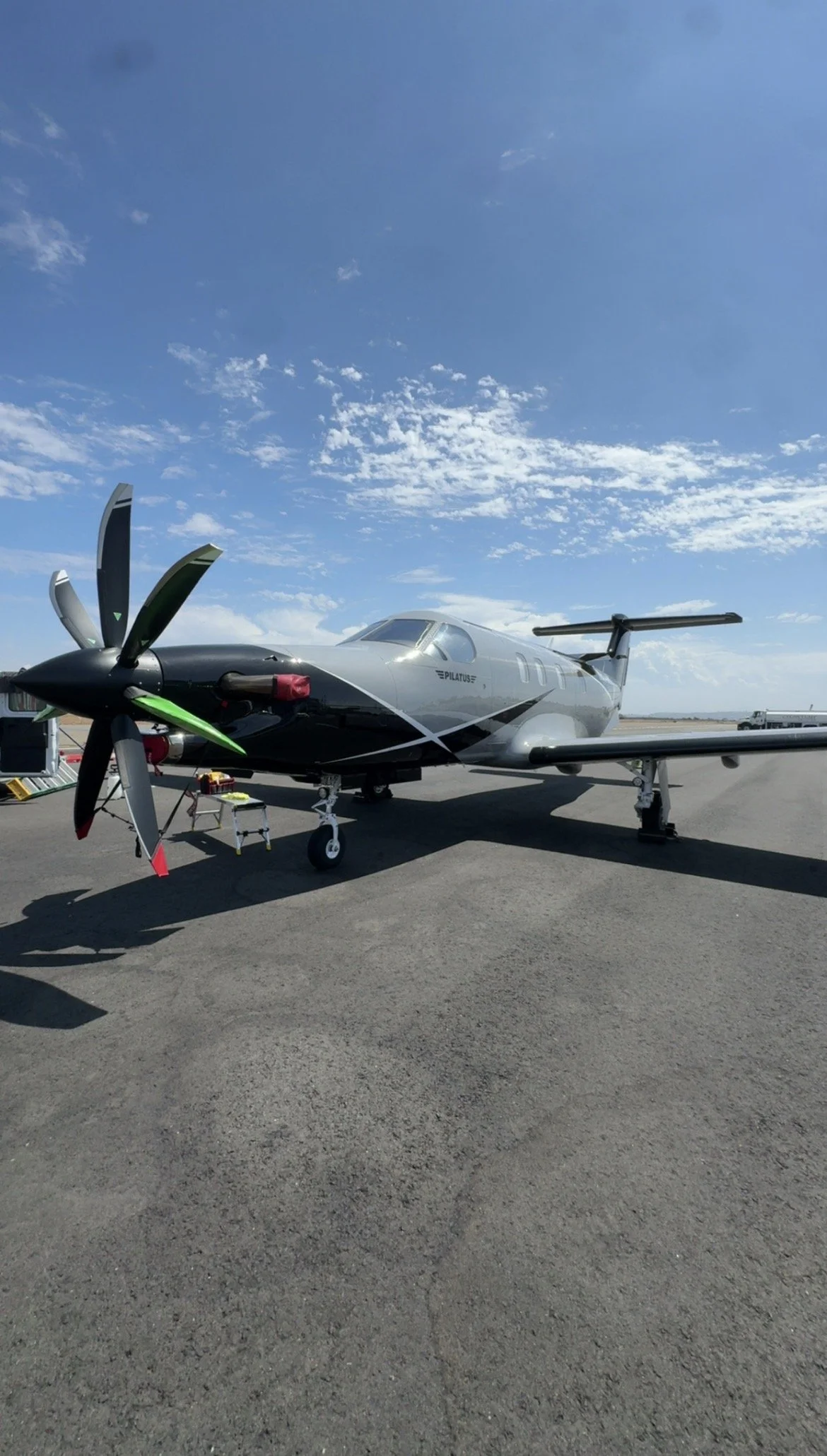 Small airplane parked on the tarmac under a blue sky with scattered clouds, with tools and equipment nearby.