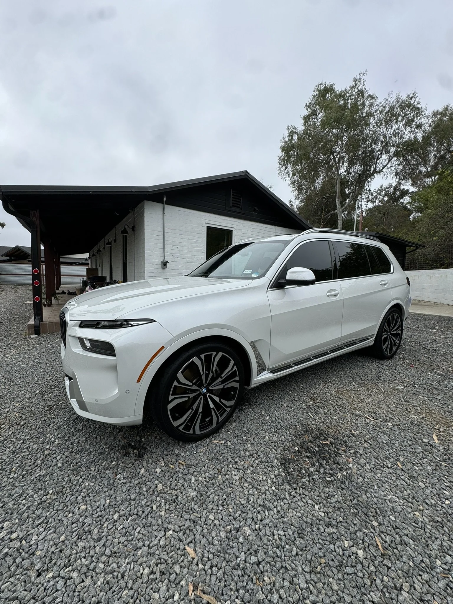 A white luxury SUV parked on a gravel driveway in front of a modern house with white brick walls and a black roof. There are trees in the background and an overcast sky above.
