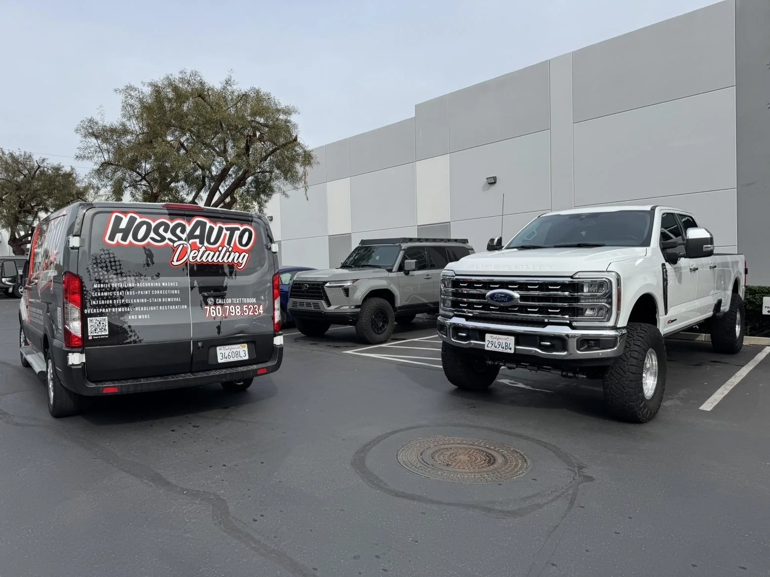 A parking lot with three vehicles: a gray van with advertising for auto detailing services, a gray SUV, and a white Ford pickup truck.