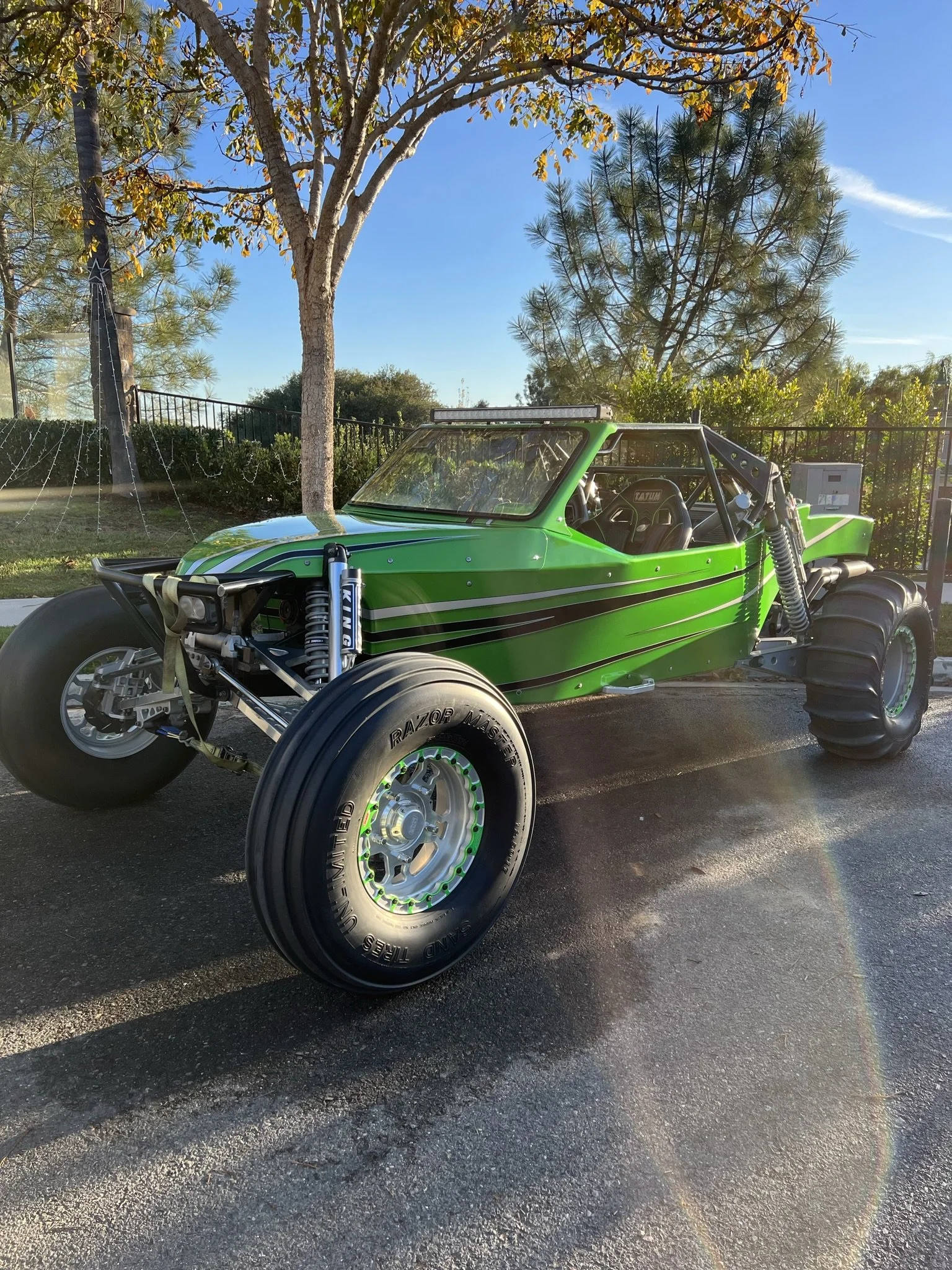 A green off-road dune buggy with black tires parked on a paved surface, trees and a fence in the background, and a clear blue sky overhead.