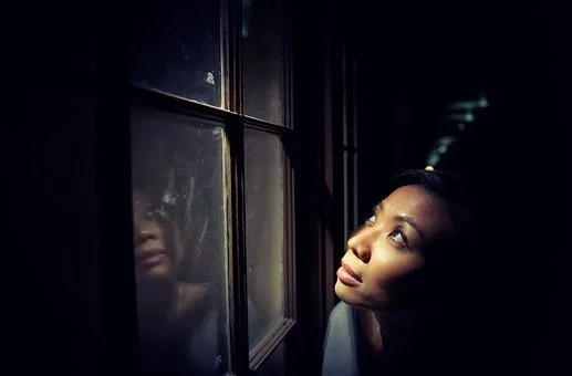 A woman looking out a window at night, with her reflection visible on the glass.