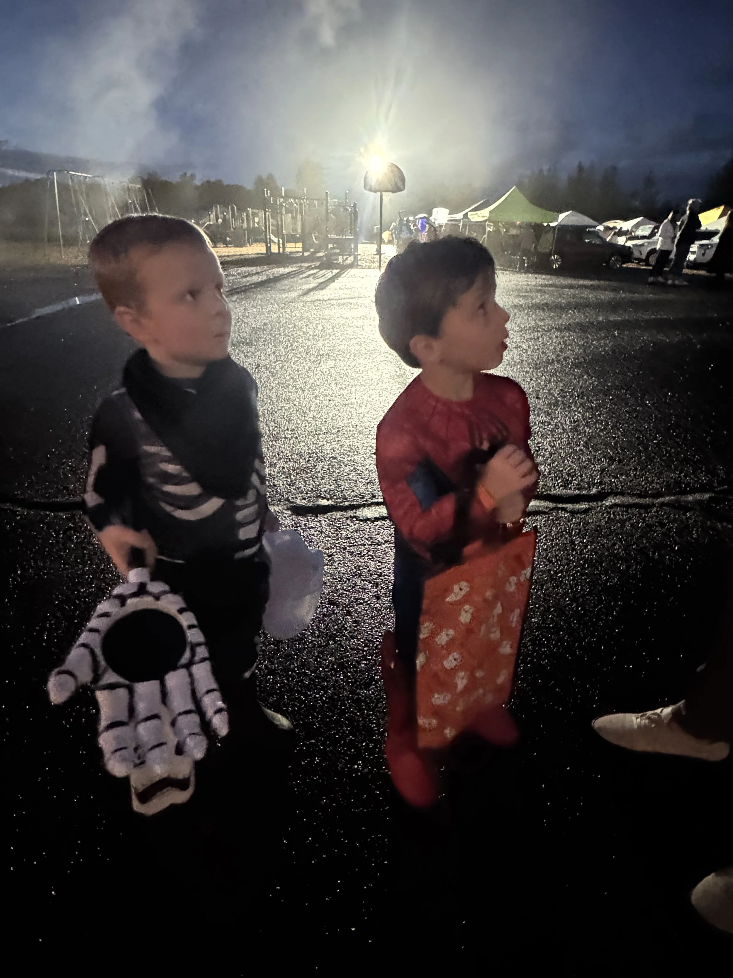Two young boys dressed in Halloween costumes standing on a wet parking lot at night, holding bags for trick-or-treating, with tents and cars in the background.