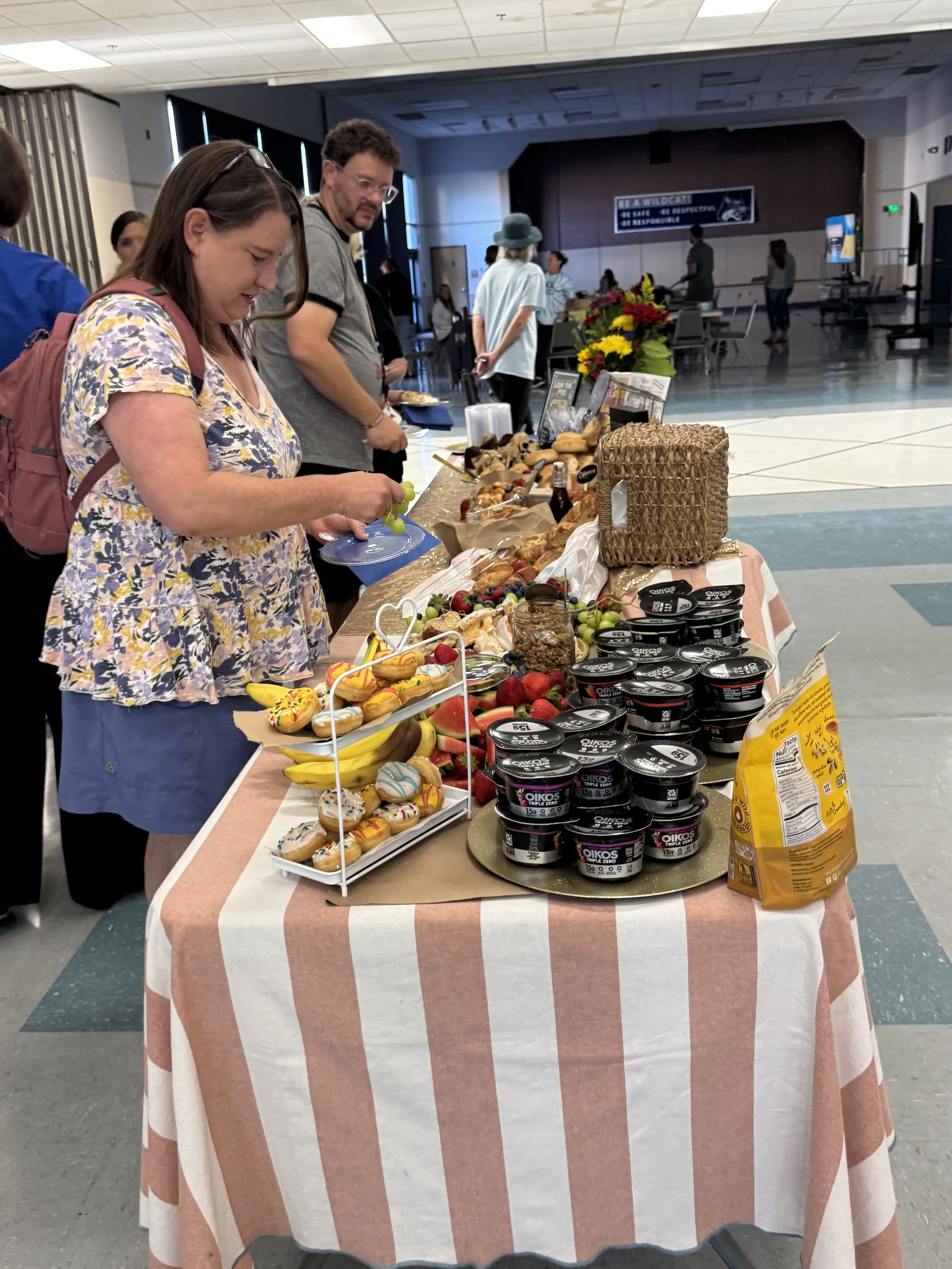 A table of snacks and desserts at an indoor event, with people browsing in the background.