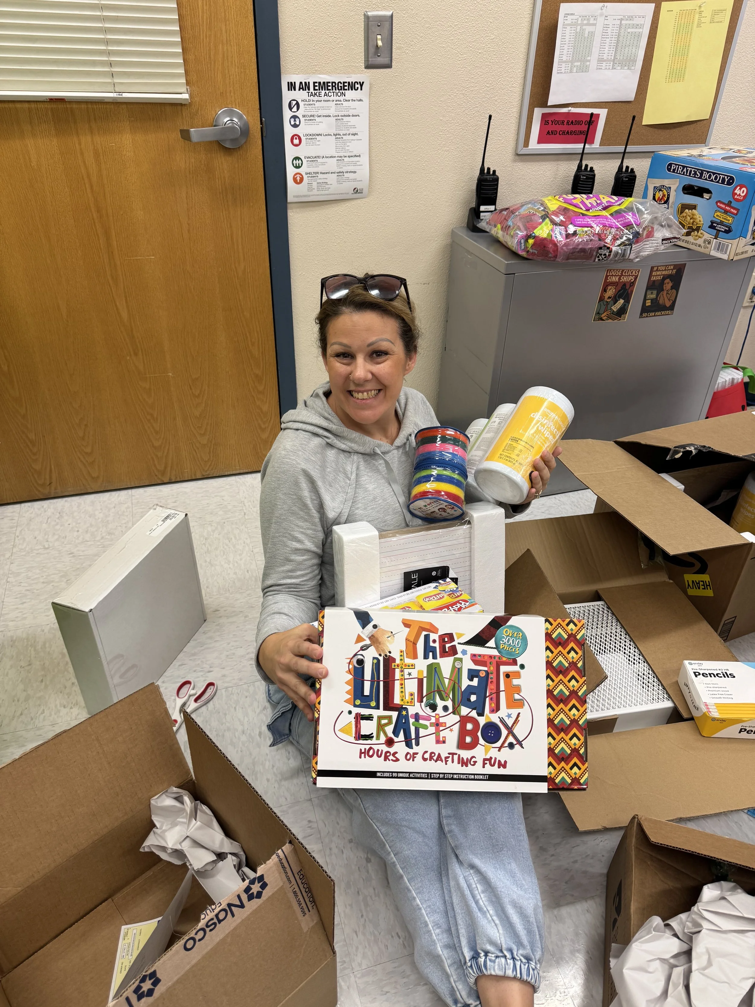 A woman sitting on the floor surrounded by cardboard boxes, holding a craft box titled 'The Ultimate Craft Box.' She is smiling and has sunglasses on her head, with crafting supplies like tape and disinfectant bottles nearby.