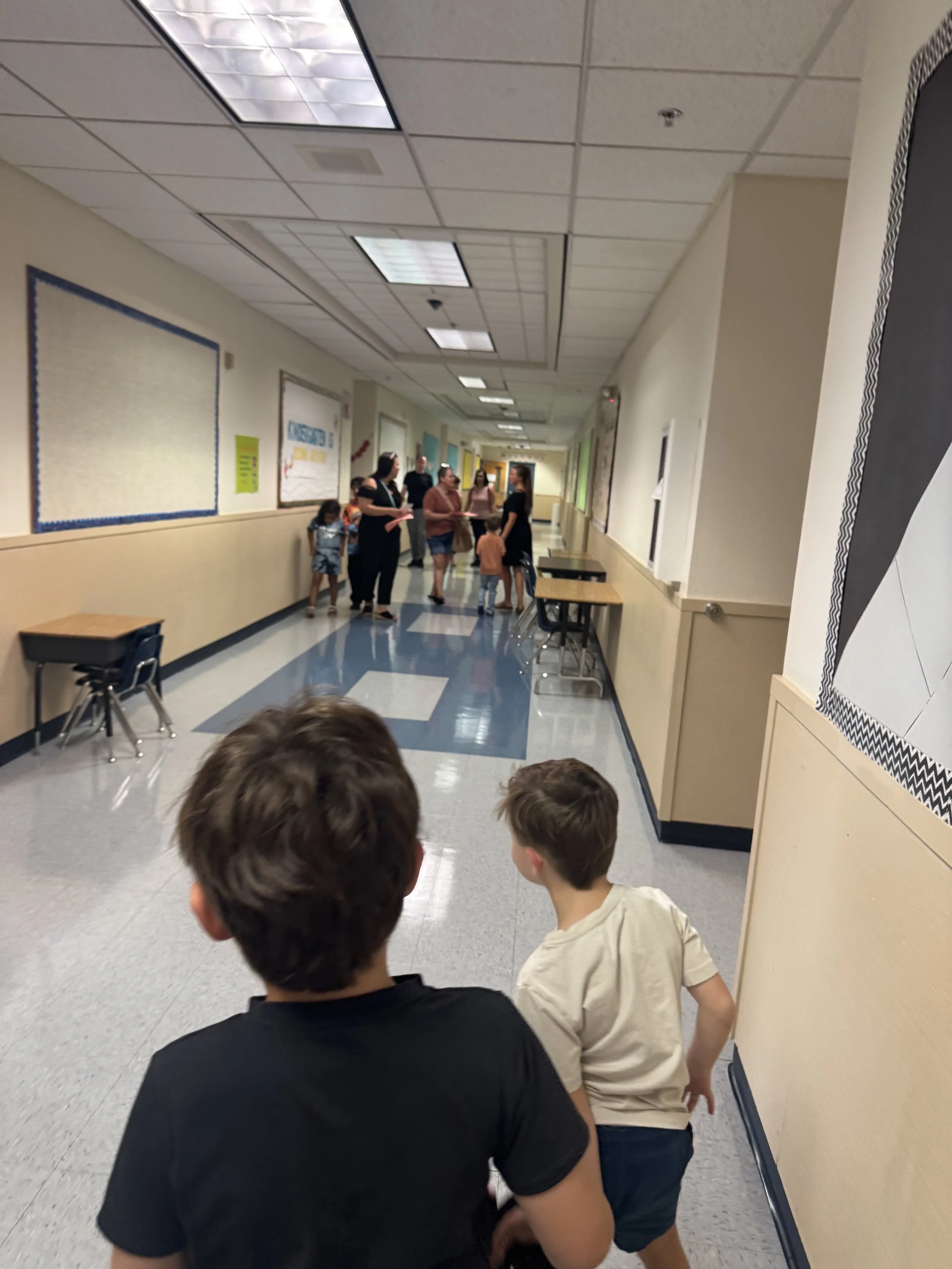 Children sitting in a school hallway watching a group of people near a bulletin board.