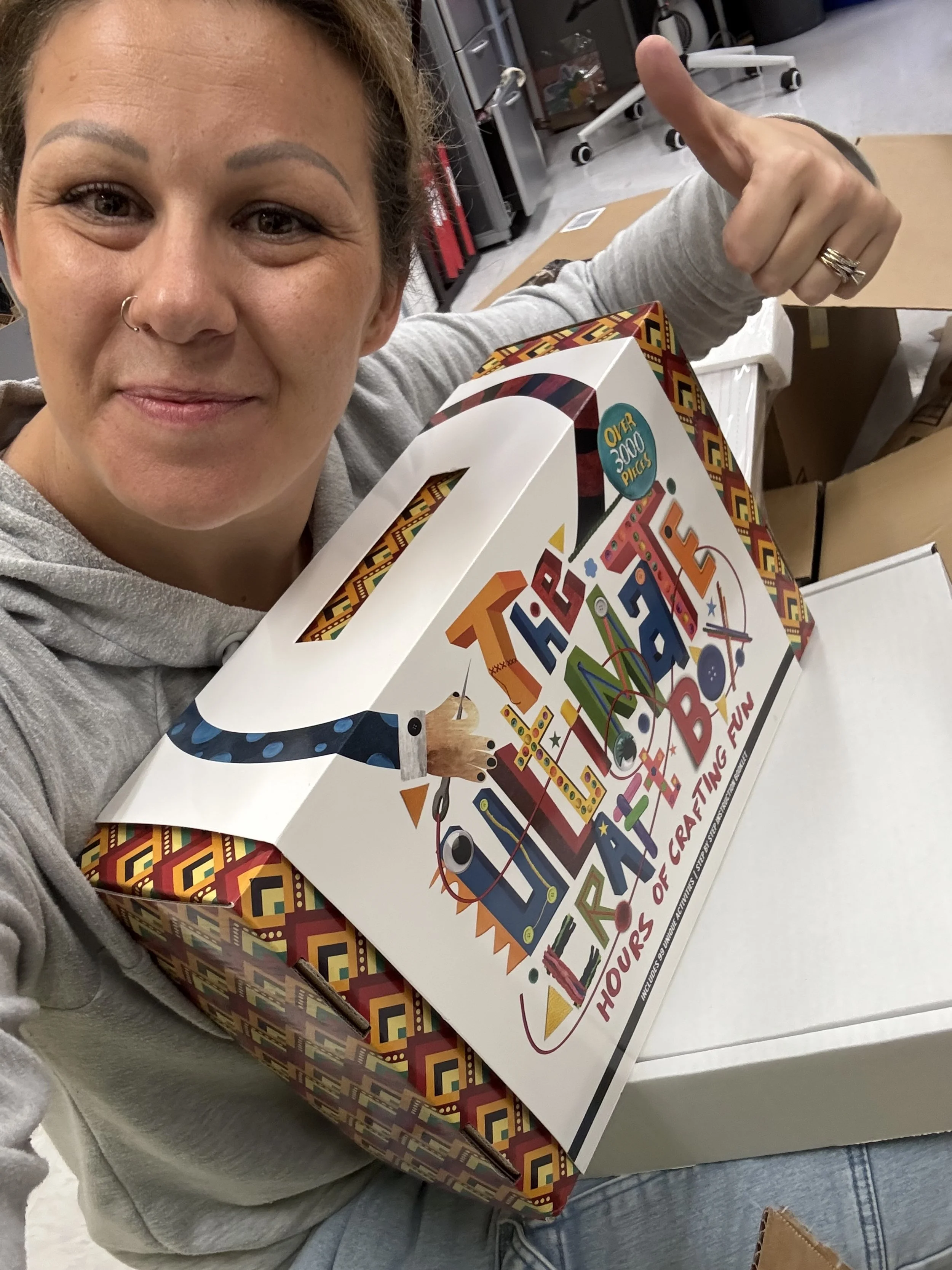Woman smiling and pointing at a colorful box labeled "60+ Hours of Creative Fun" while sitting among open boxes in a storage or warehouse space.