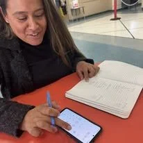Woman sitting at a table using a smartphone with notebooks open in front of her in an indoor setting, possibly an airport or train station.