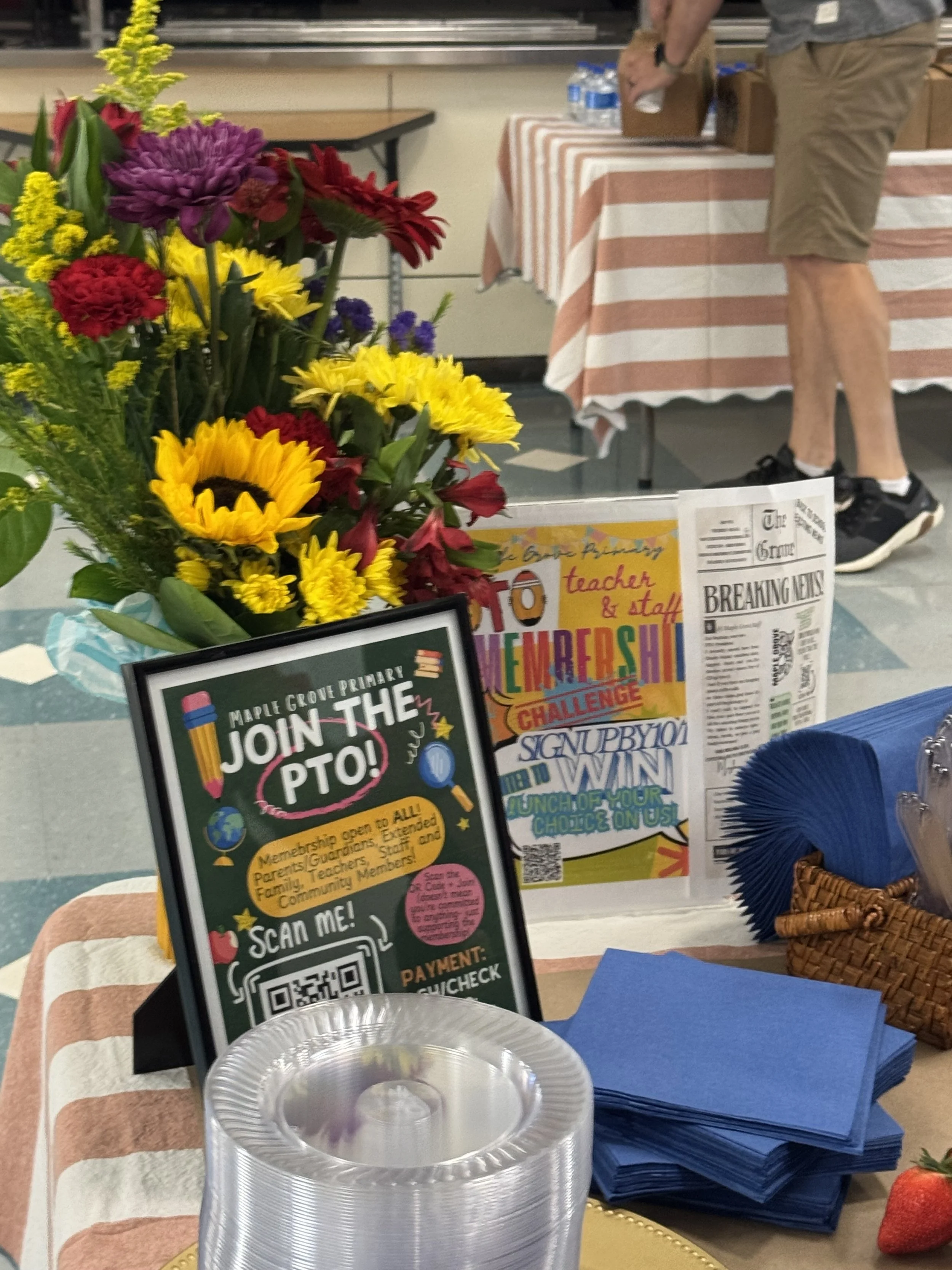 A table decorated with a colorful flower arrangement and various printed signs promoting the PTO membership, a teacher and staff appreciation challenge, and a community event. Blue napkins and a stack of clear plastic cups are also on the table.
