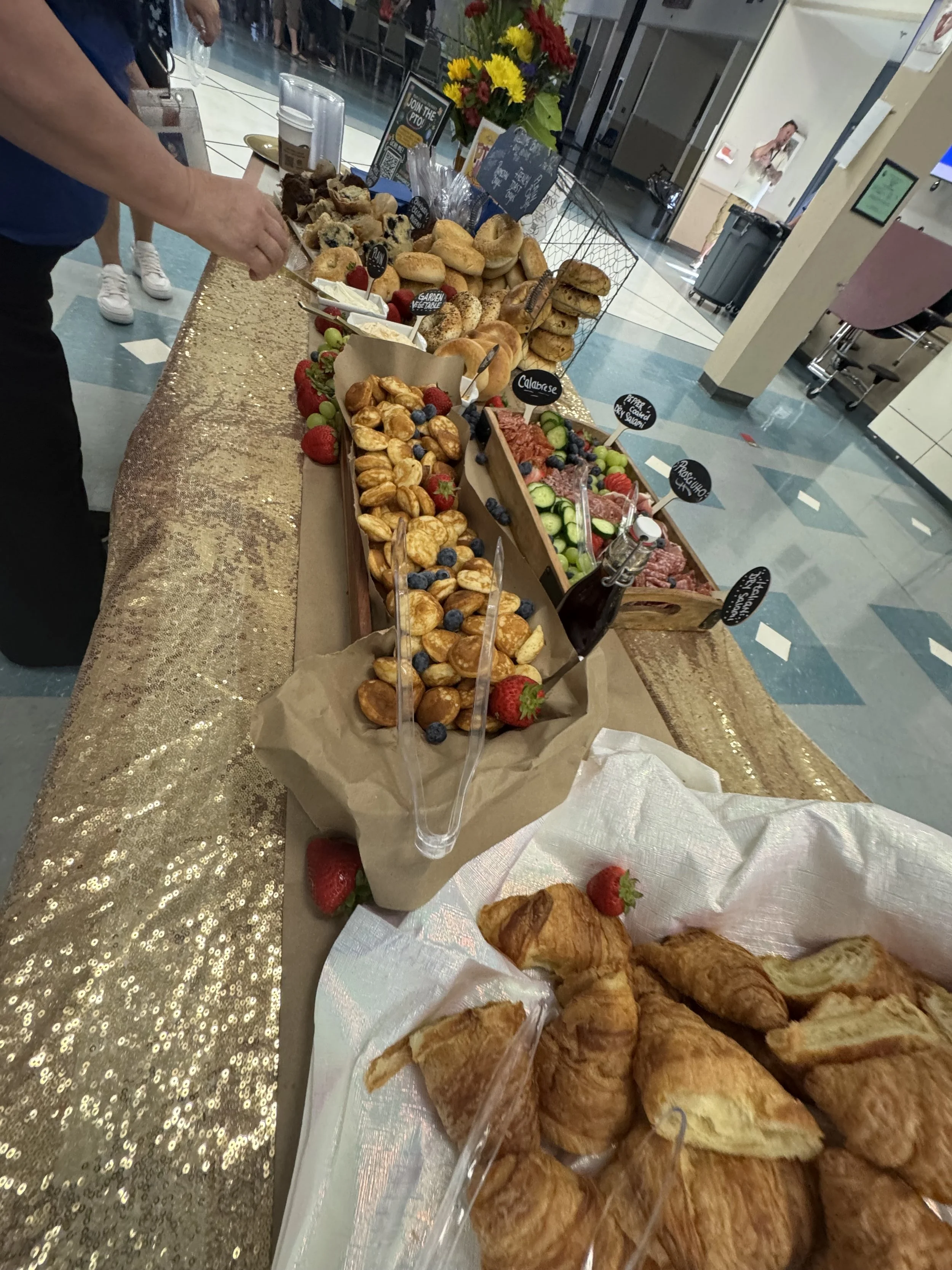A breakfast buffet with croissants, fresh fruit, bagels, muffins, and pastries on a gold sequin tablecloth in a public space.