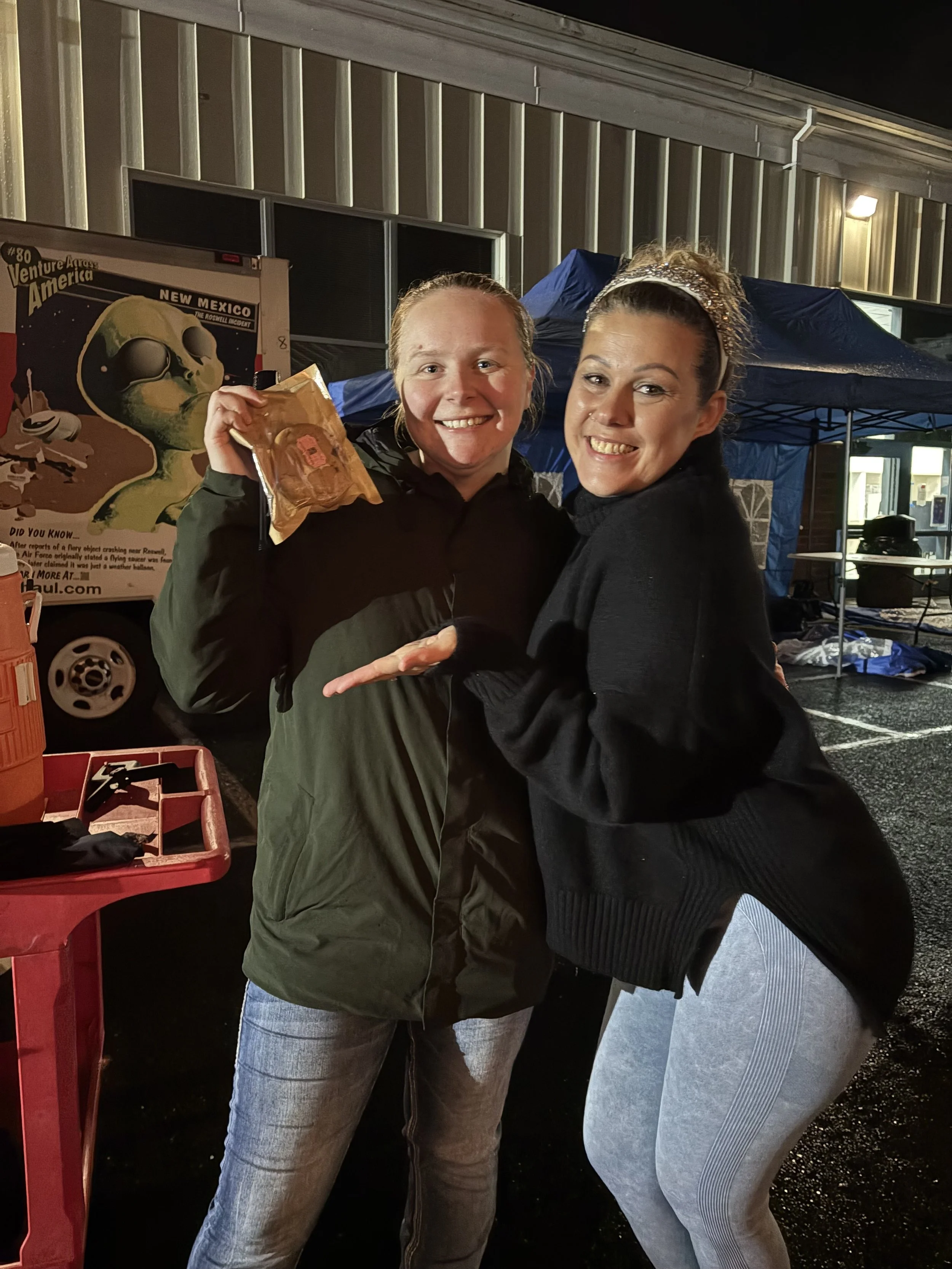 Two women smiling at night outdoors, one holding a snack bag, the other with hands out. Background includes a poster, a blue tent, and a building with lights.