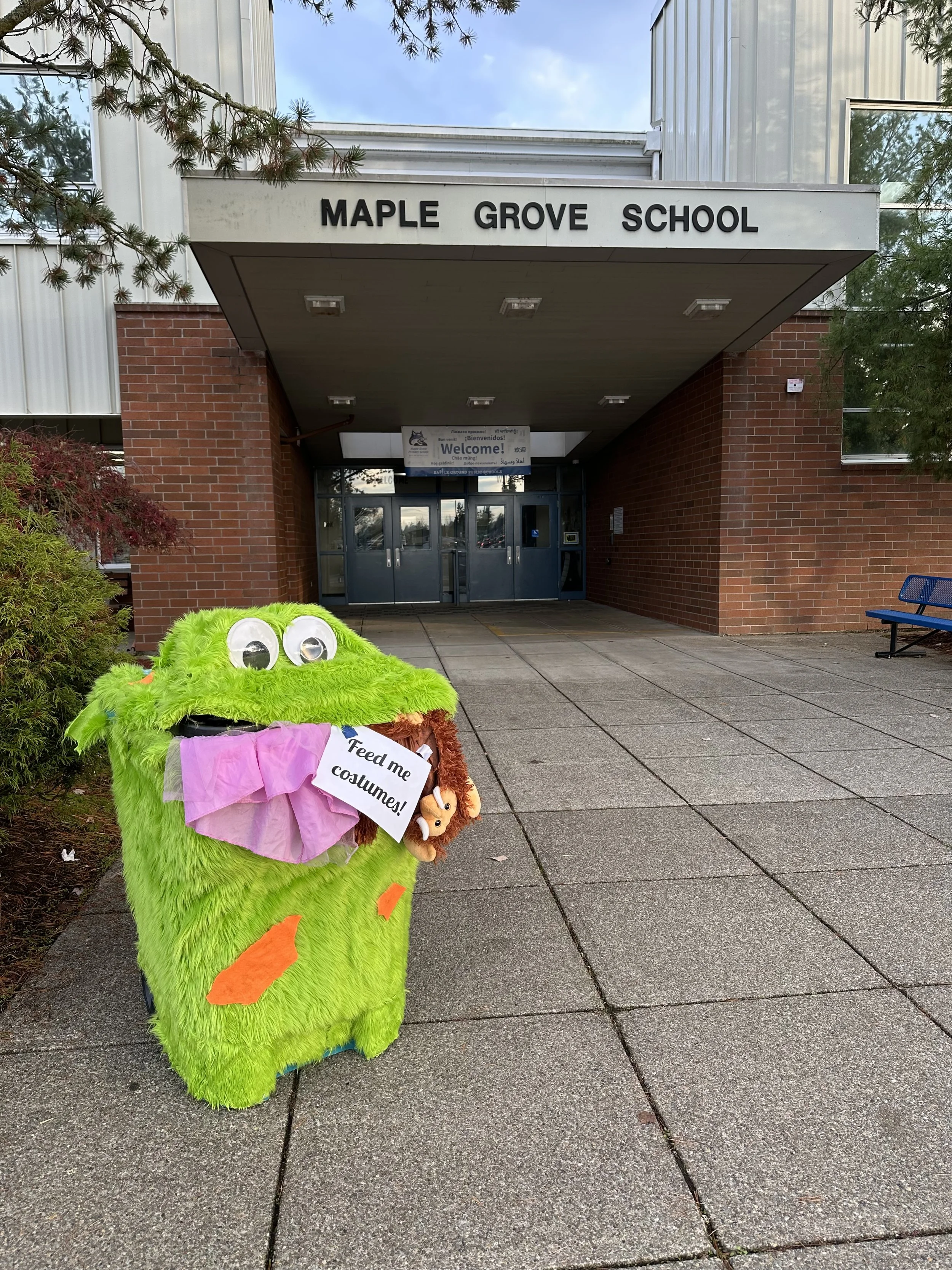 Colorful plush costume of a green monster with googly eyes, a pink tissue paper tongue, and a sign that says "Feed me costumes!" is placed outside Maple Grove School entrance on a concrete sidewalk with brick walls, trees, and glass doors in the back