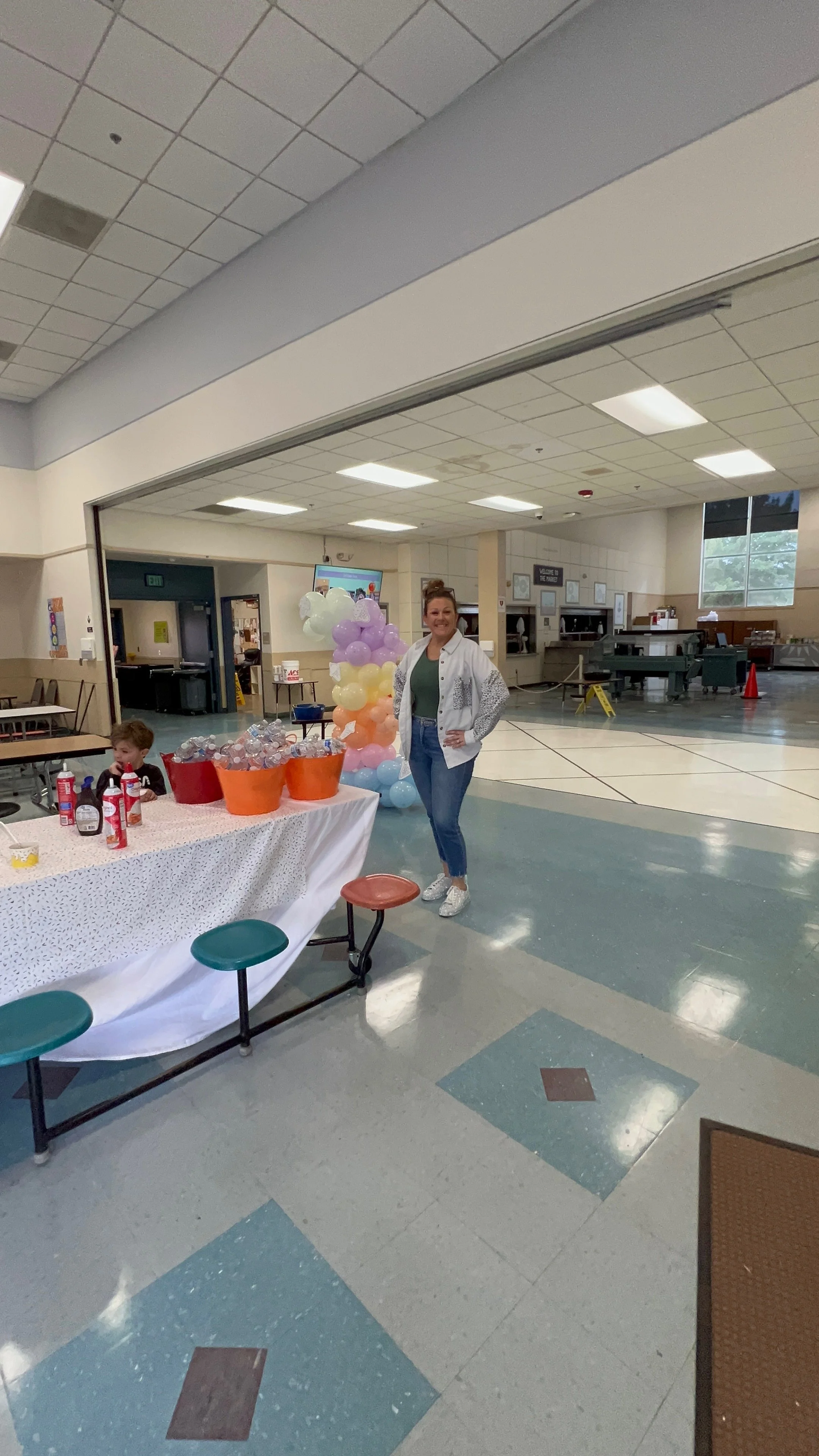 A woman standing next to a table decorated with balloons and party supplies in a room, possibly a community center or school gymnasium.