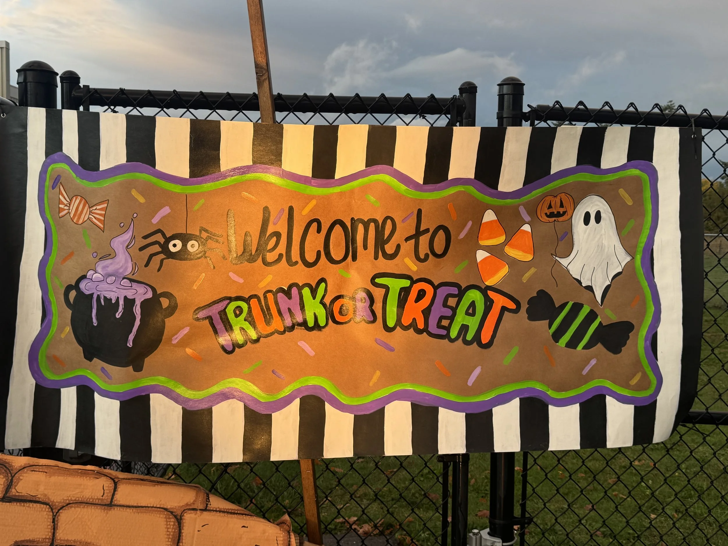 Colorful Halloween-themed banner with striped black and white border, decorated with illustrations of a purple bubbling cauldron, spider, witch hat, ghost, candy, and pumpkin. The banner reads: "Welcome to Trunk or Treat" with festive orange, green, 