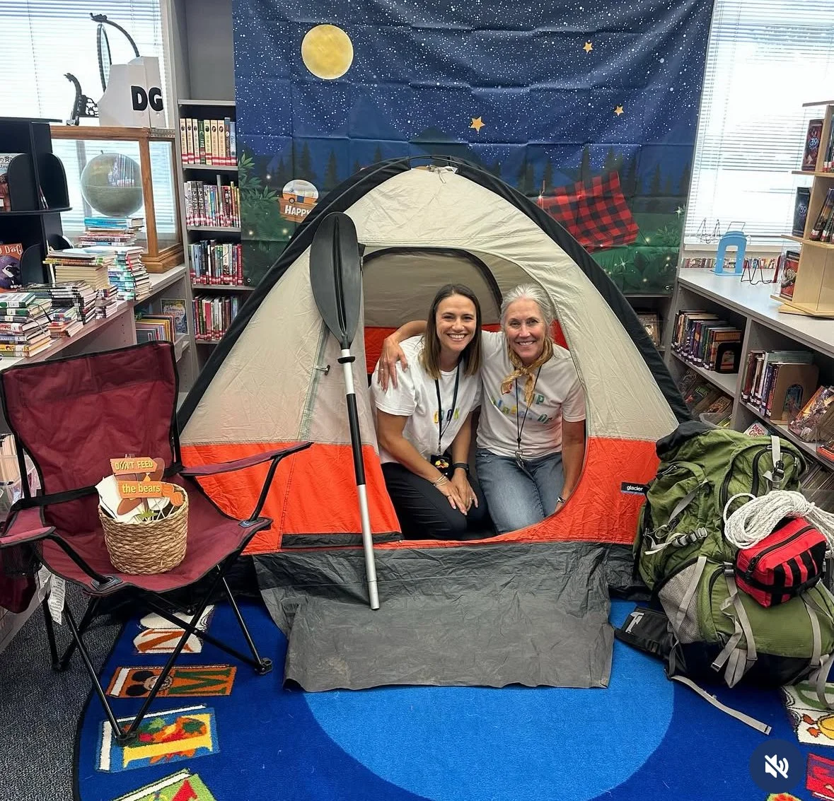Two women smiling inside a camping tent set up indoors in a library, surrounded by bookshelves, with a starry night and moon mural on the wall behind them, and camping gear like backpacks, a chair, and paddles around them.