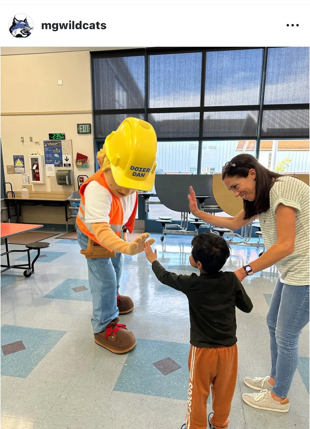 A woman and two children in an indoor setting, with the woman and a mascot giving high fives. The mascot is dressed as a construction worker with a yellow hard hat labeled "DOZER DAN," oversized shoes, and a safety vest. The children are reaching out