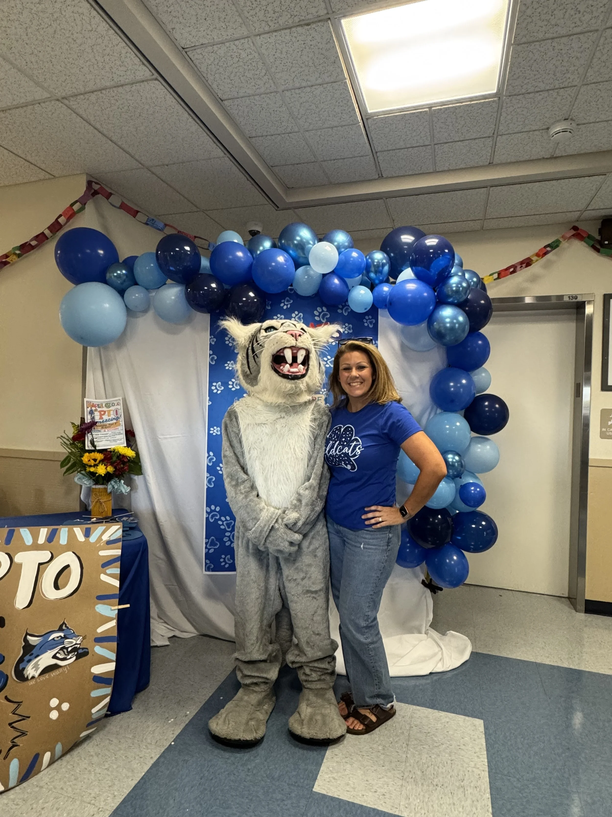 A woman in a blue T-shirt and jeans standing next to a person dressed in a wildcat mascot costume, both smiling, in front of a backdrop with blue balloons and paw print decorations, at a school event.