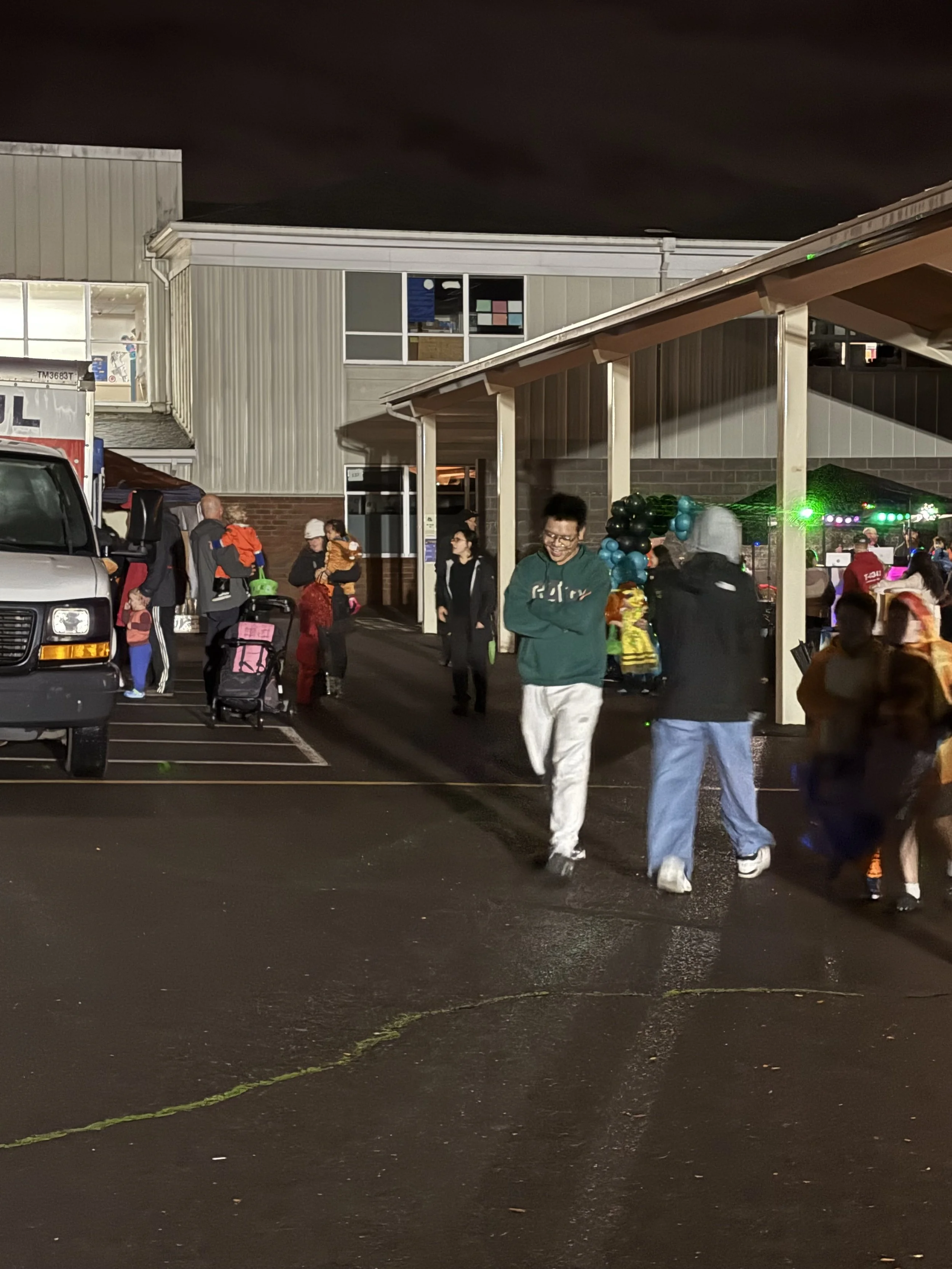 Nighttime scene outside a building with people walking and gathering, some carrying children, with balloons and decorations, and a truck parked nearby.