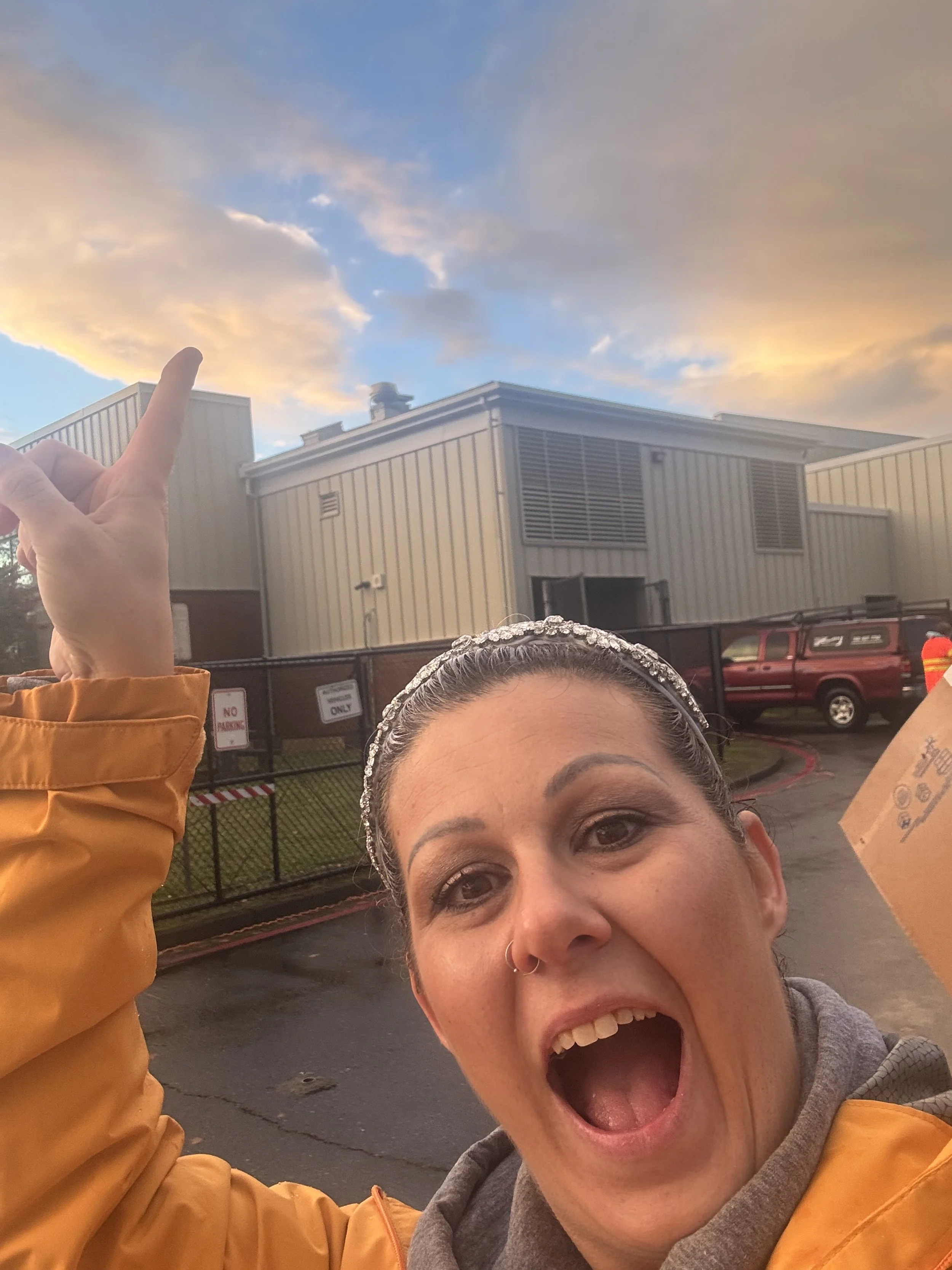 Close-up of a woman with a surprised expression, wearing a gray hoodie and a shiny headband, holding a cardboard box, standing outside in front of a fence and a beige industrial building, with a colorful cloudy sky in the background.