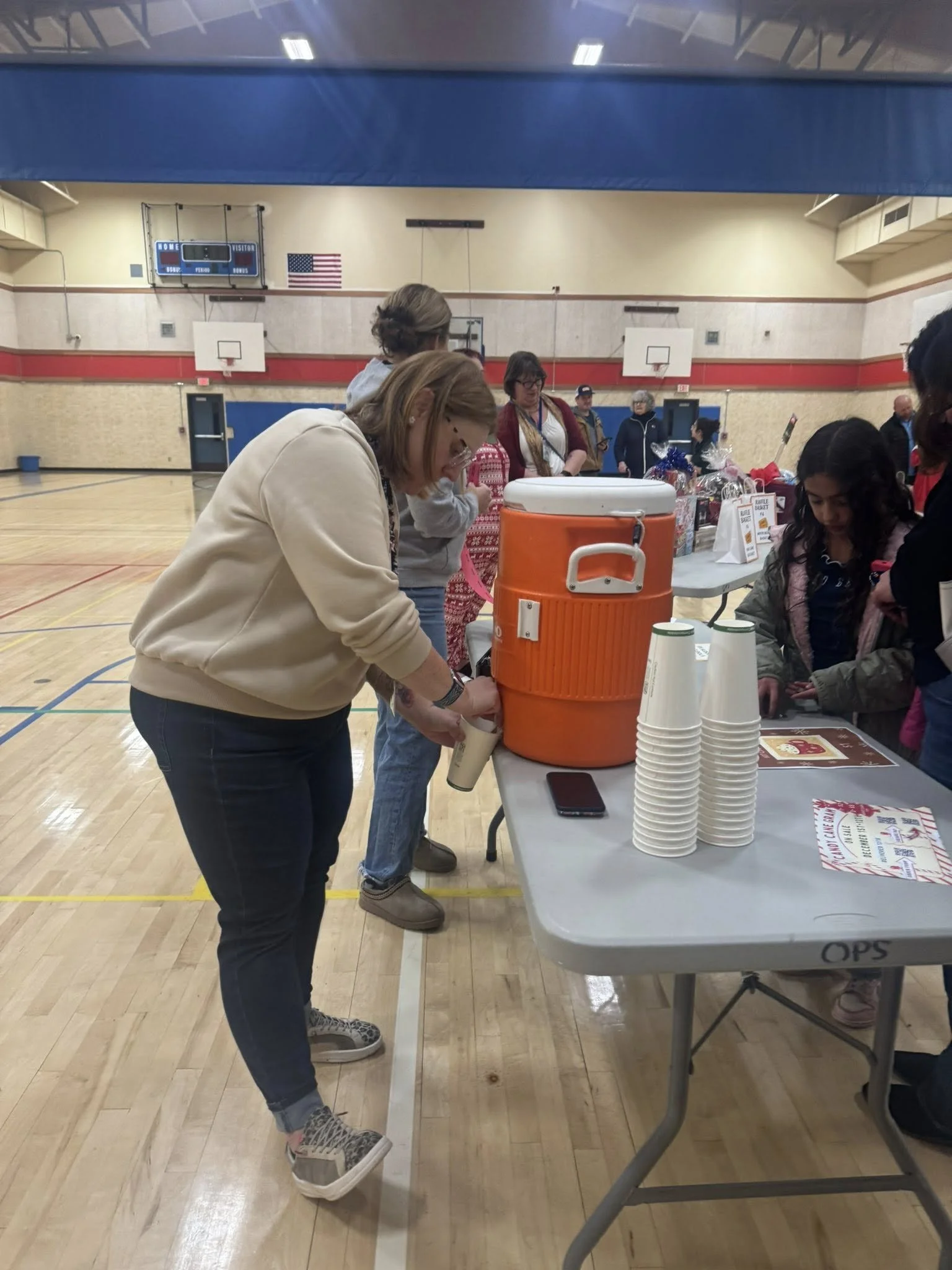 People gathered around a table in a gymnasium with a large orange cooler, paper cups, and a phone on the table, likely at a community event or fundraiser.