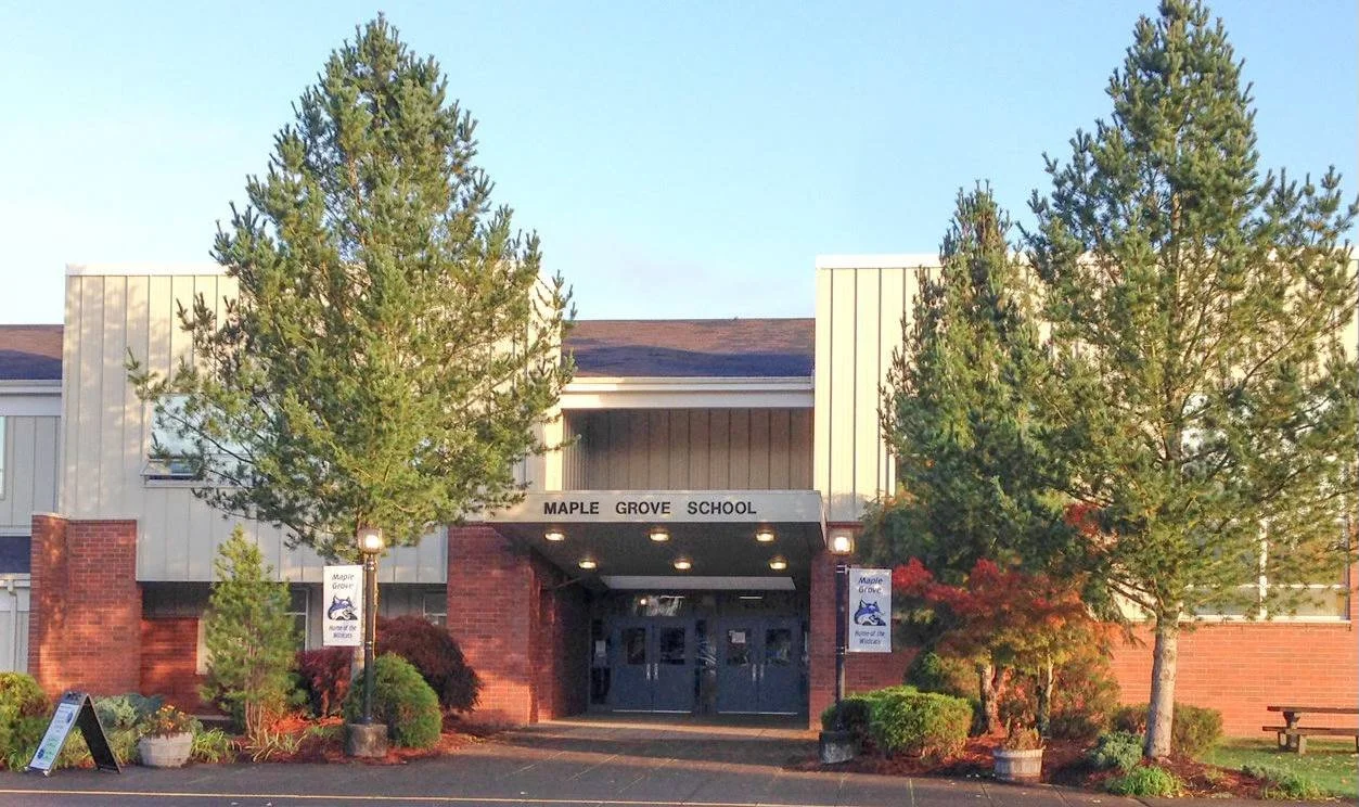 Front view of Maple Grove School, surrounded by trees and bushes, with a brick and metal building exterior and signs on either side of the entrance.