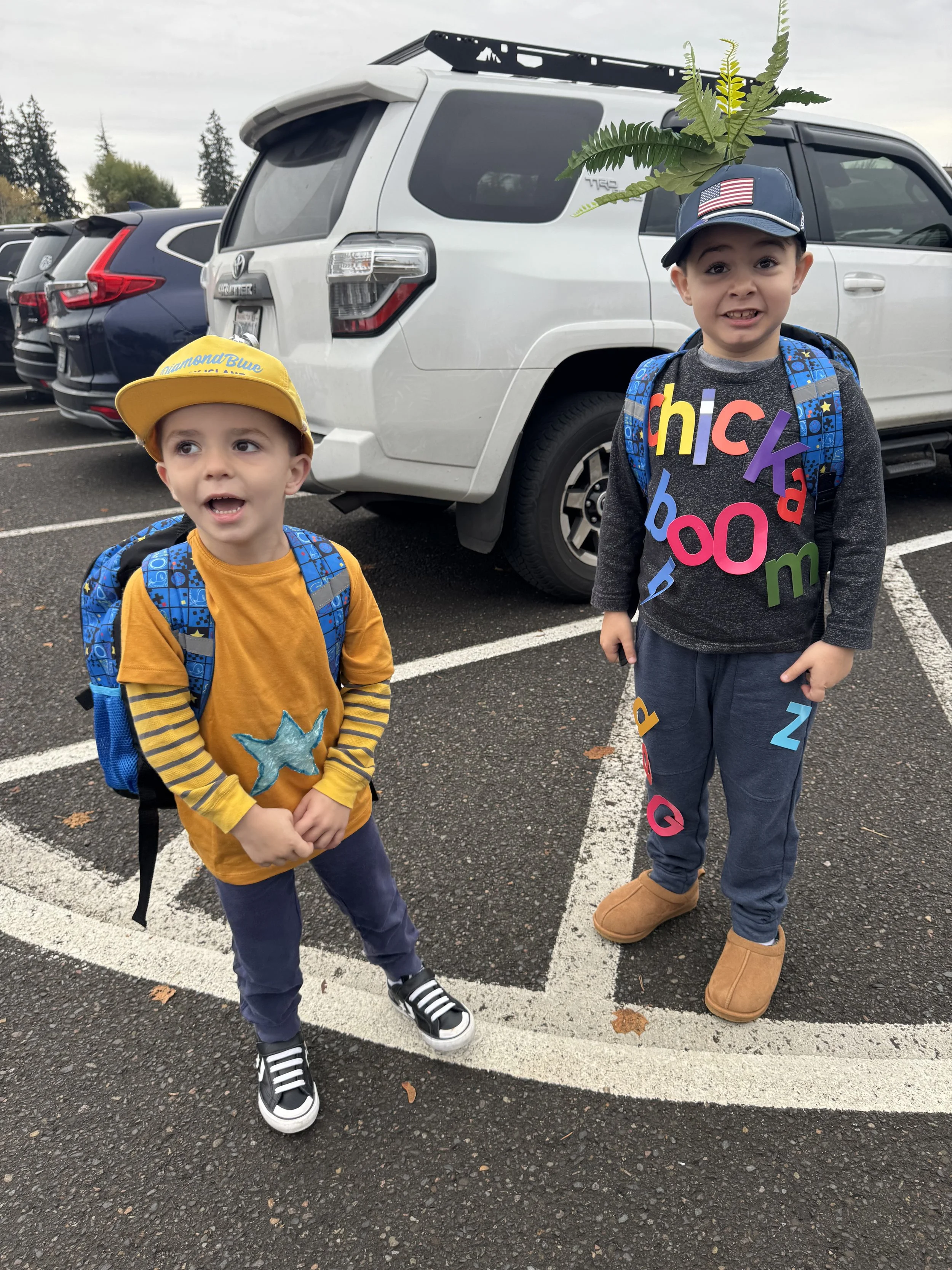 Two young boys standing in a parking lot next to a white SUV, wearing backpacks and hats, one with a sunny yellow cap and the other with a navy blue cap. The boy on the right has a shirt with colorful letters, and the boy on the left has a yellow shi