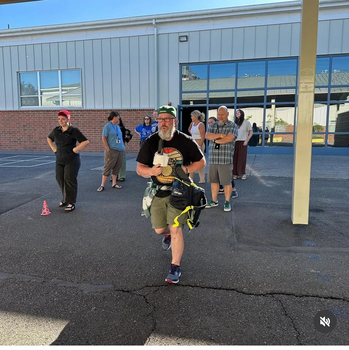 A man running outdoors, holding a microphone, with a group of people standing behind him near a building with large windows. The man has a beard, glasses, and wears a green cap and casual clothing. The group appears to be waiting or observing.