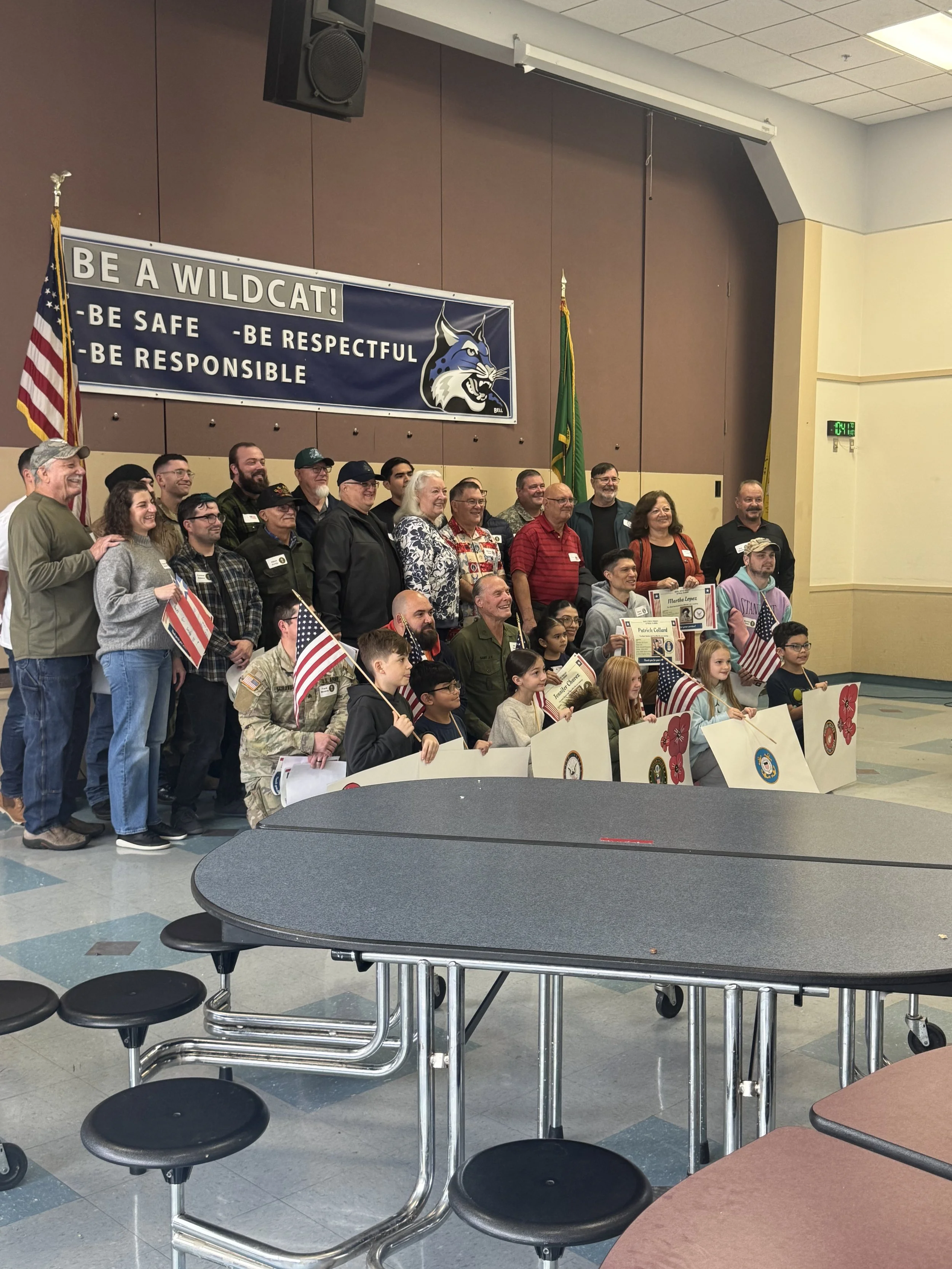 A group of adults and children gathered on a stage for a patriotic or military recognition event, holding American flags and certificates. The backdrop features a large banner with the phrase "Be a Wildcat!" and safety and respect qualities, with a w