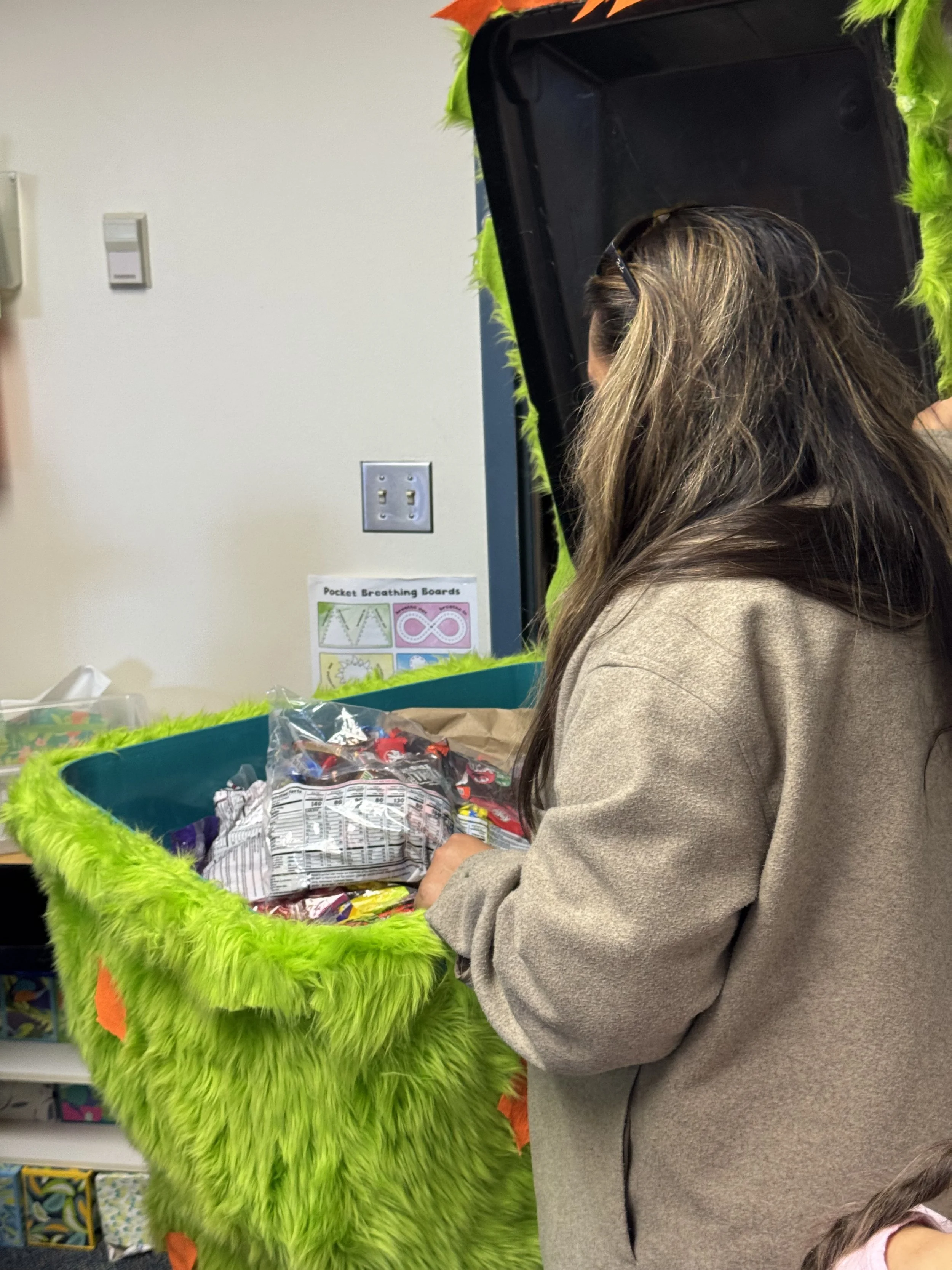 A woman with long brown hair wearing a beige jacket is standing at a table with a large green furry container filled with snacks. She is looking into or reaching into the container, which has assorted packaged snacks inside.