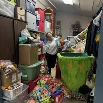 A child in a laundry room surrounded by clutter, with laundry, storage bins, and various household items visible.