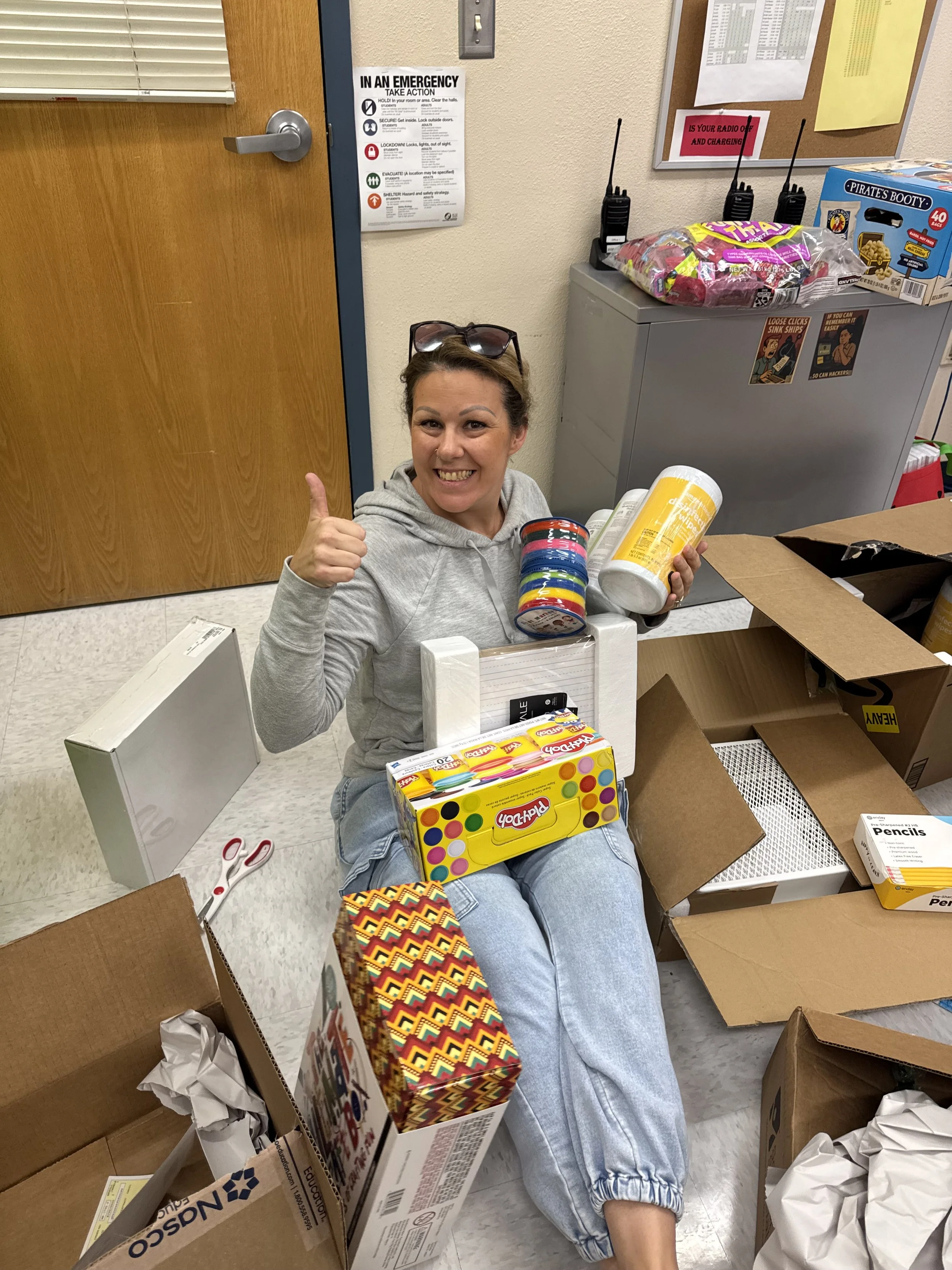 A woman sitting on the floor surrounded by cardboard boxes, holding a box of Play-Doh, with a big smile and giving a thumbs-up.