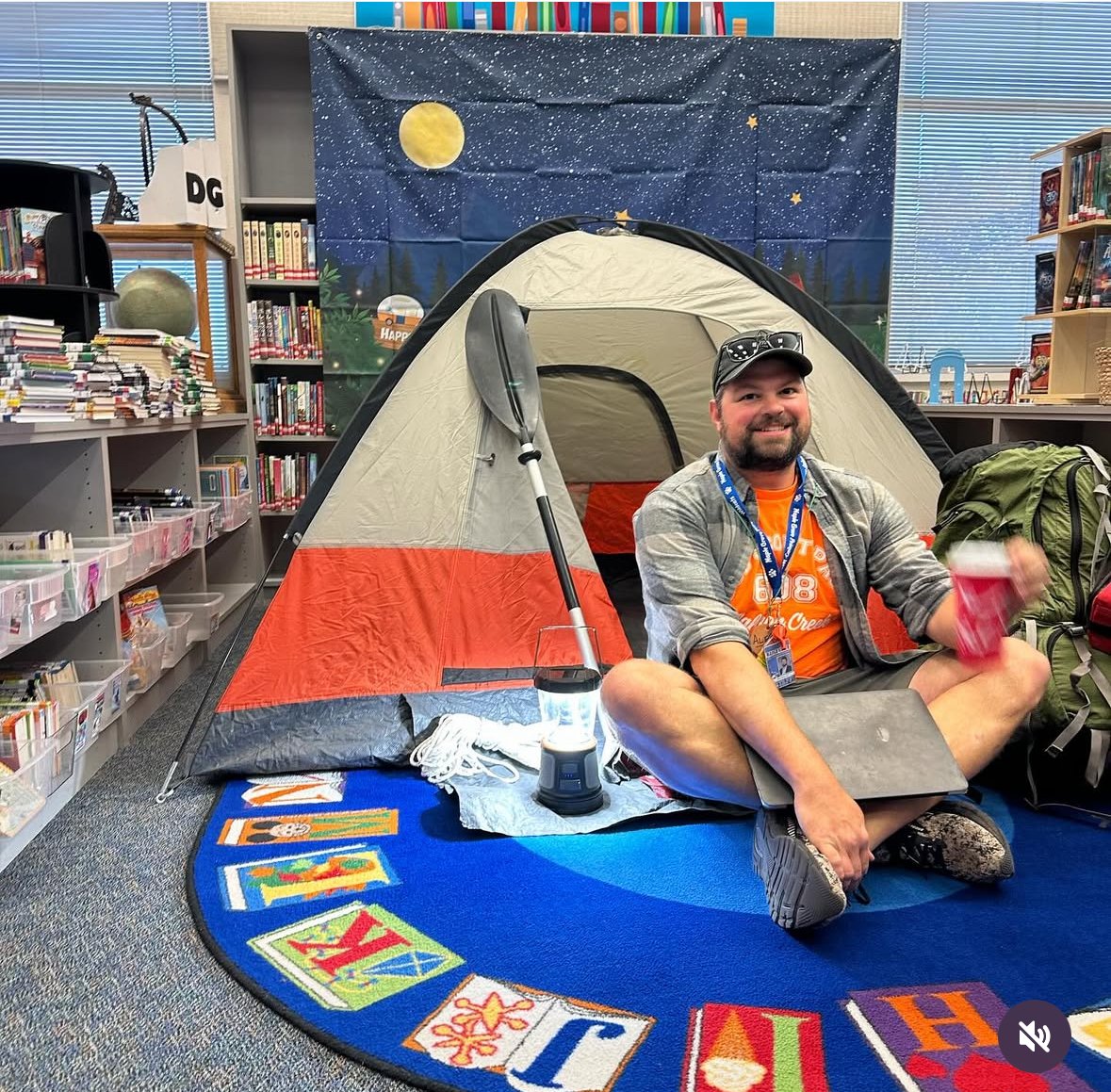 A man sitting on a colorful alphabet-themed rug inside a library, in front of a camping tent with a backdrop of stars, a moon, and trees. He is smiling, wearing a cap, sunglasses, and an orange T-shirt, and has a backpack beside him.