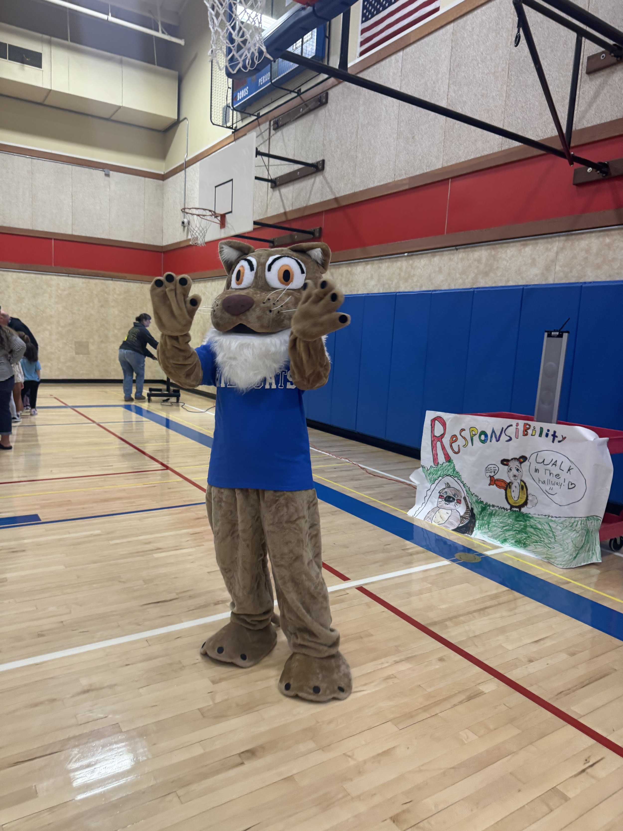 Person dressed in a lion mascot costume wearing a blue shirt, standing in a gymnasium with a basketball hoop, and a colorful sign that says 'Responsibility' in the background.