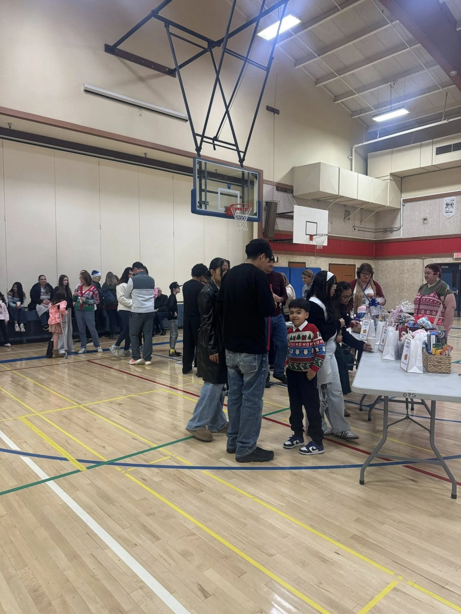 People standing in line at a holiday-themed event inside a gymnasium, with a table of gift bags and holiday decorations.