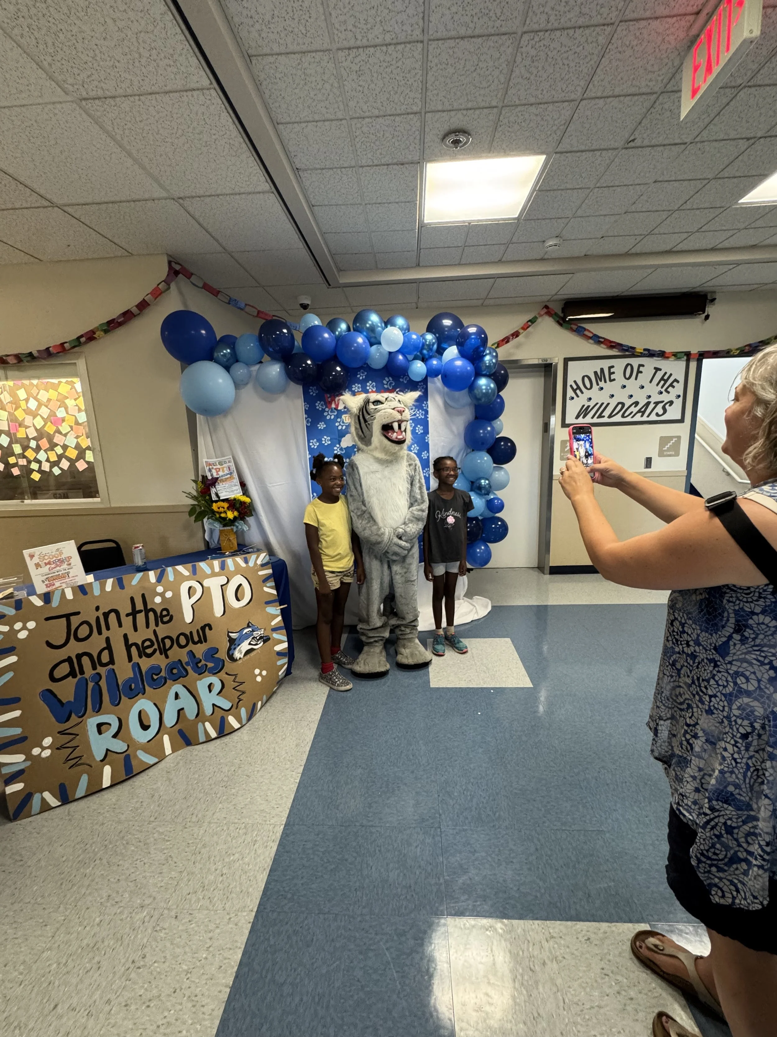 Two children and a person in a wildcat mascot costume posing for a photo at a school event with balloons and a sign that reads 'Join the PTO and help our Wildcats ROAR'.
