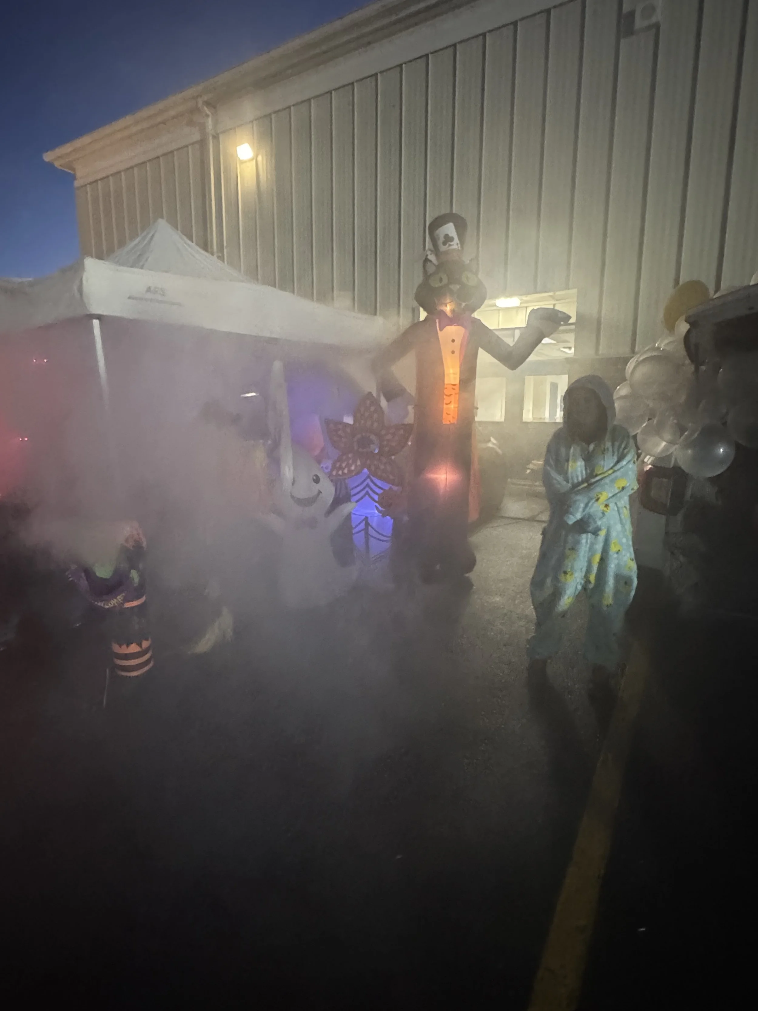 A Halloween party scene outdoors with fog. A person in a clown costume with a glowing orange chest, wearing a tall black hat, stands next to a young girl in pajamas, both near a decorated booth with balloons, a smiling ghost figure, and Halloween-the
