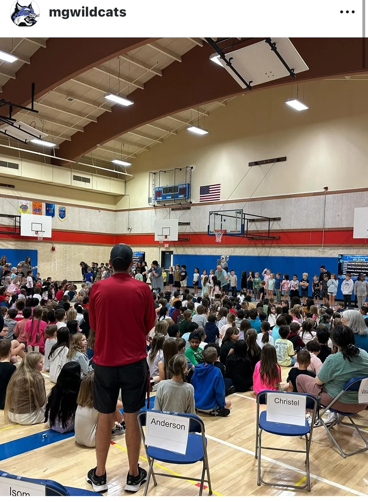 A school gymnasium filled with students sitting on the floor and on chairs, and children on stage during a school event with a mascot and teachers.
