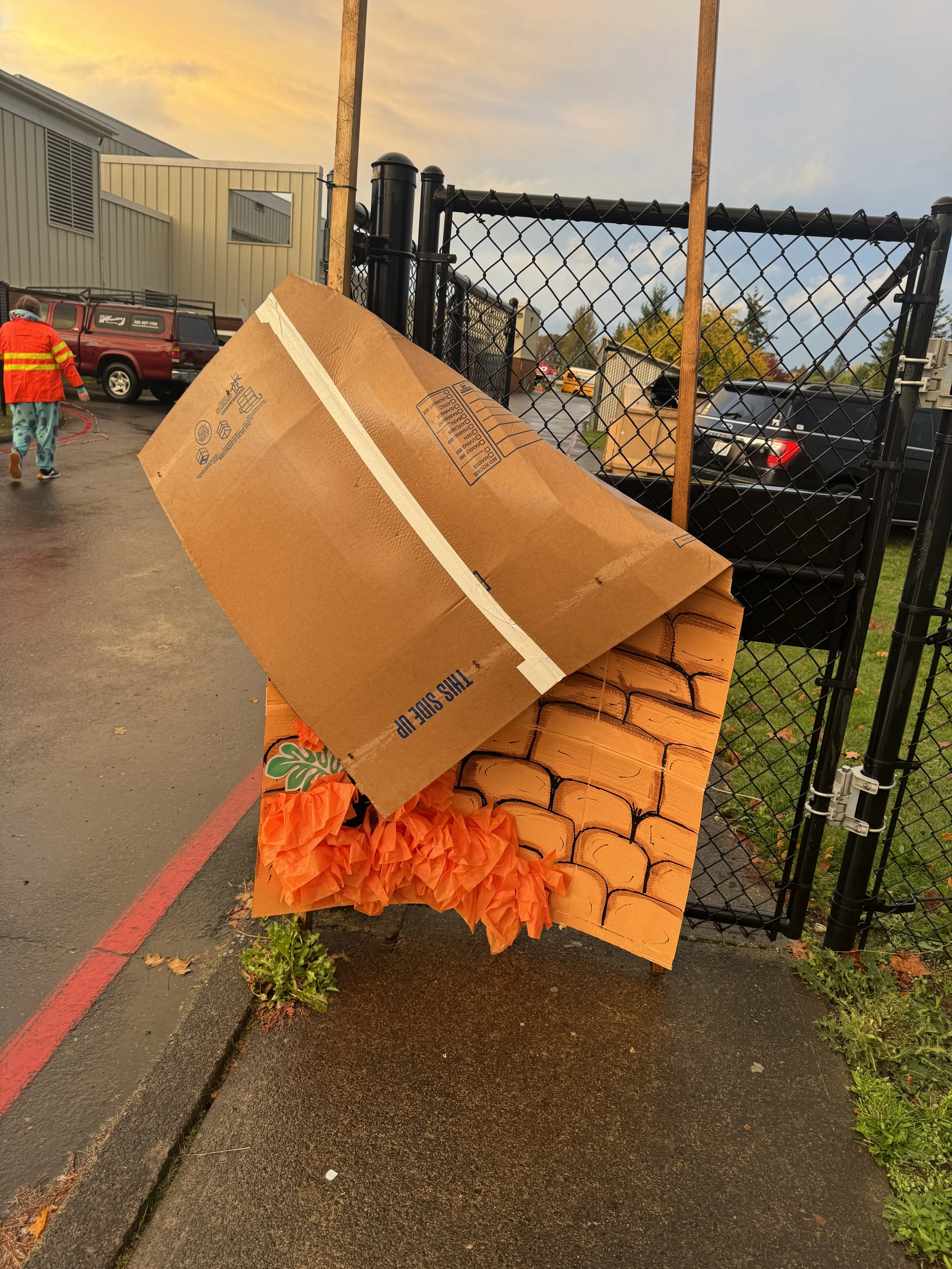 Decorative cardboard display resembling a brick wall with orange paper flames, positioned next to a chain-link fence outside during sunset.