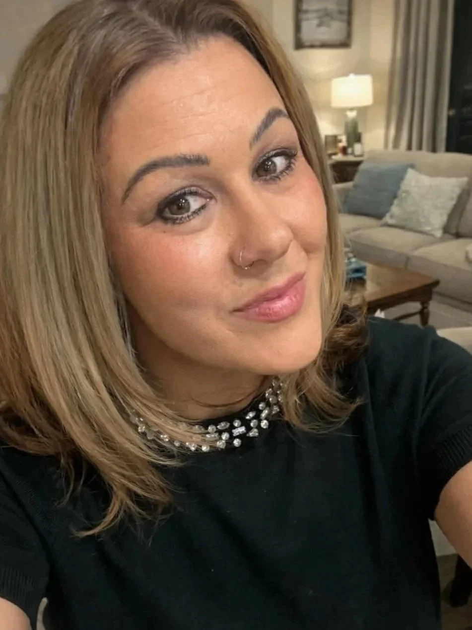 A woman with shoulder-length blonde hair, wearing a black top and a pearl necklace, taking a selfie in a living room with a beige sofa, pillows, a wooden coffee table, and a lamp in the background.