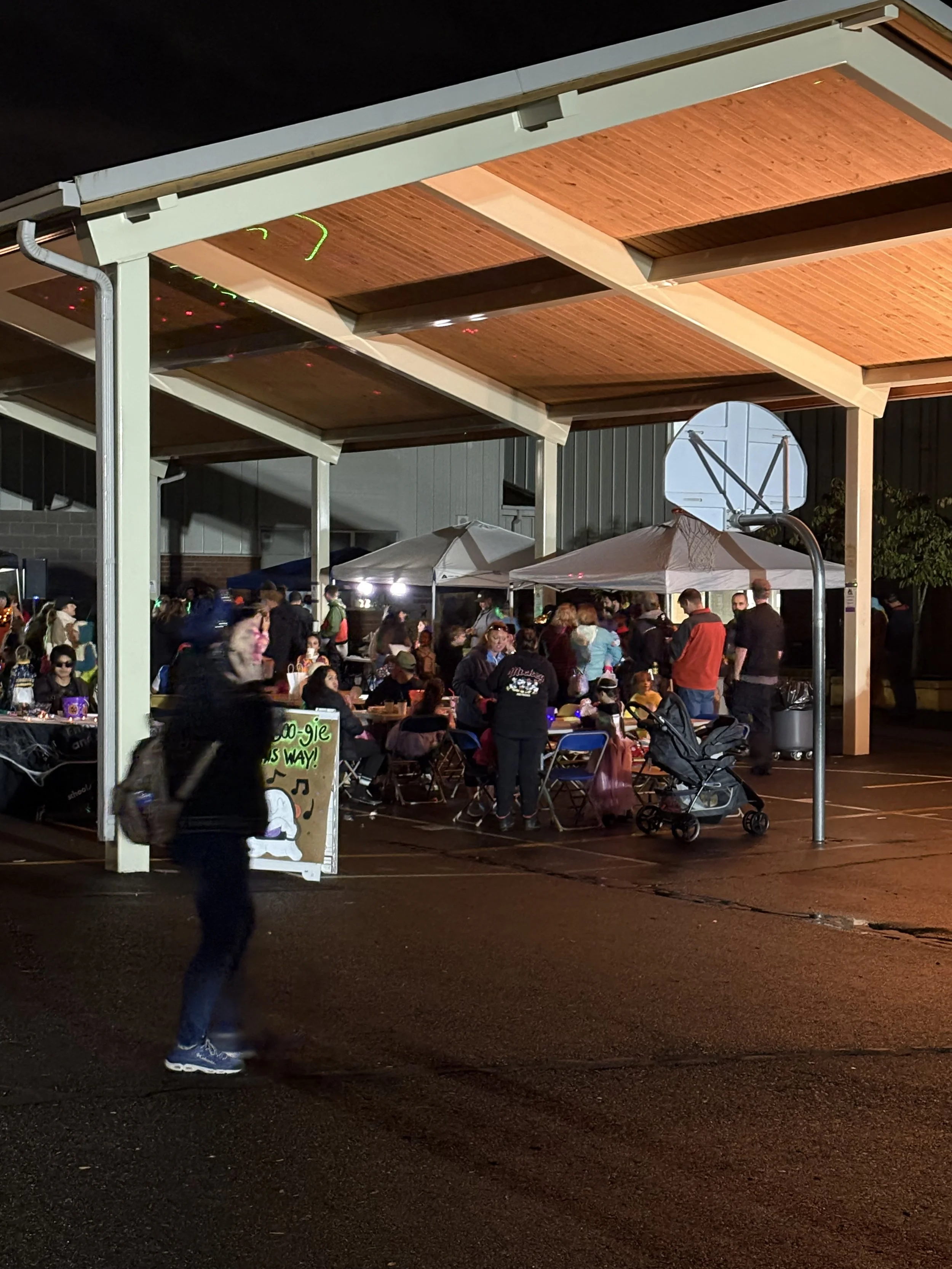 Nighttime outdoor gathering with people around tables under a covered pavilion, food and drinks being served, and a person walking past with a stroller nearby.