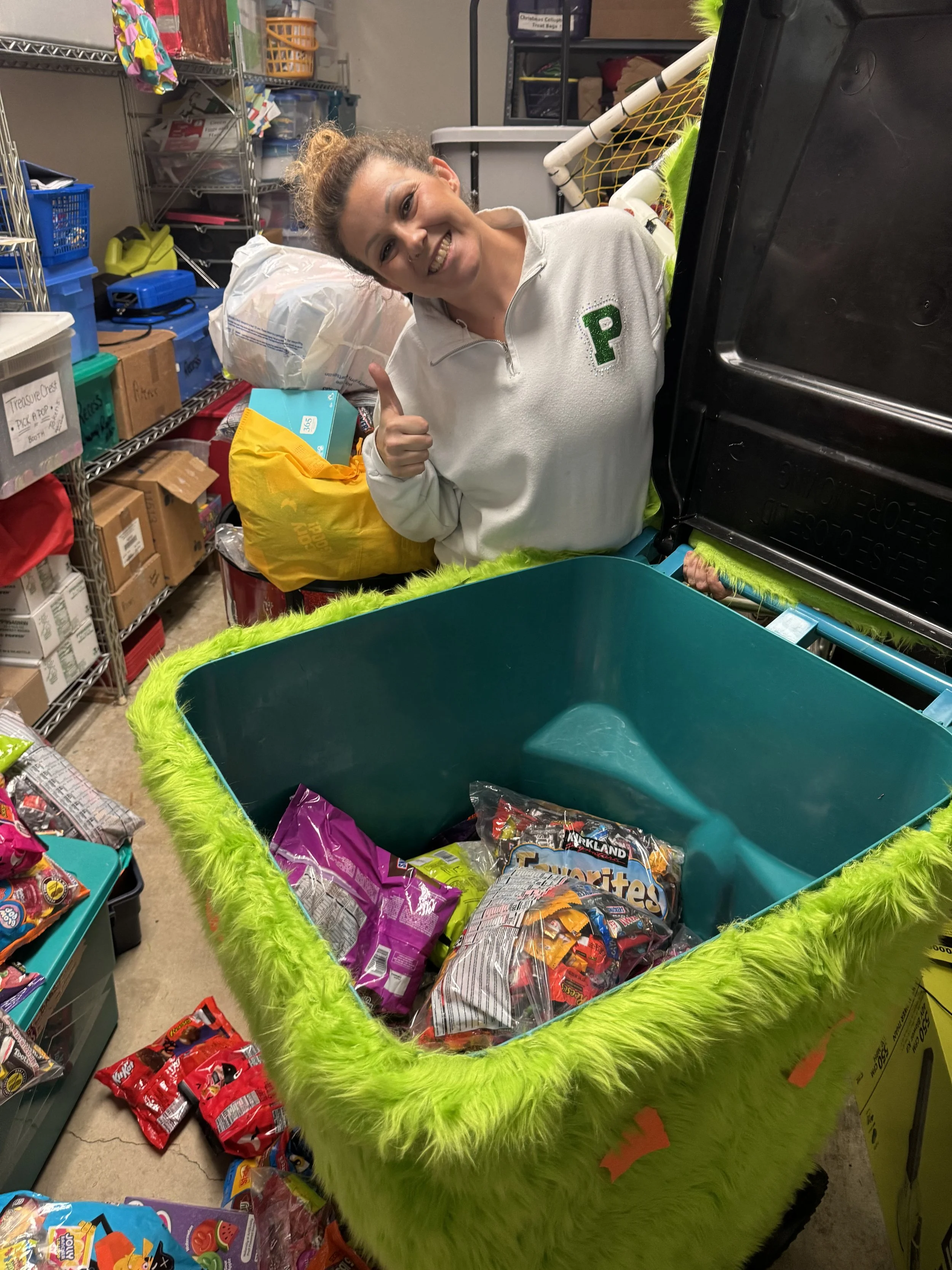 A woman smiling and giving a thumbs-up while leaning over a large, green, furry candy collector filled with assorted bags of candy, in a storage room with shelves and boxes.
