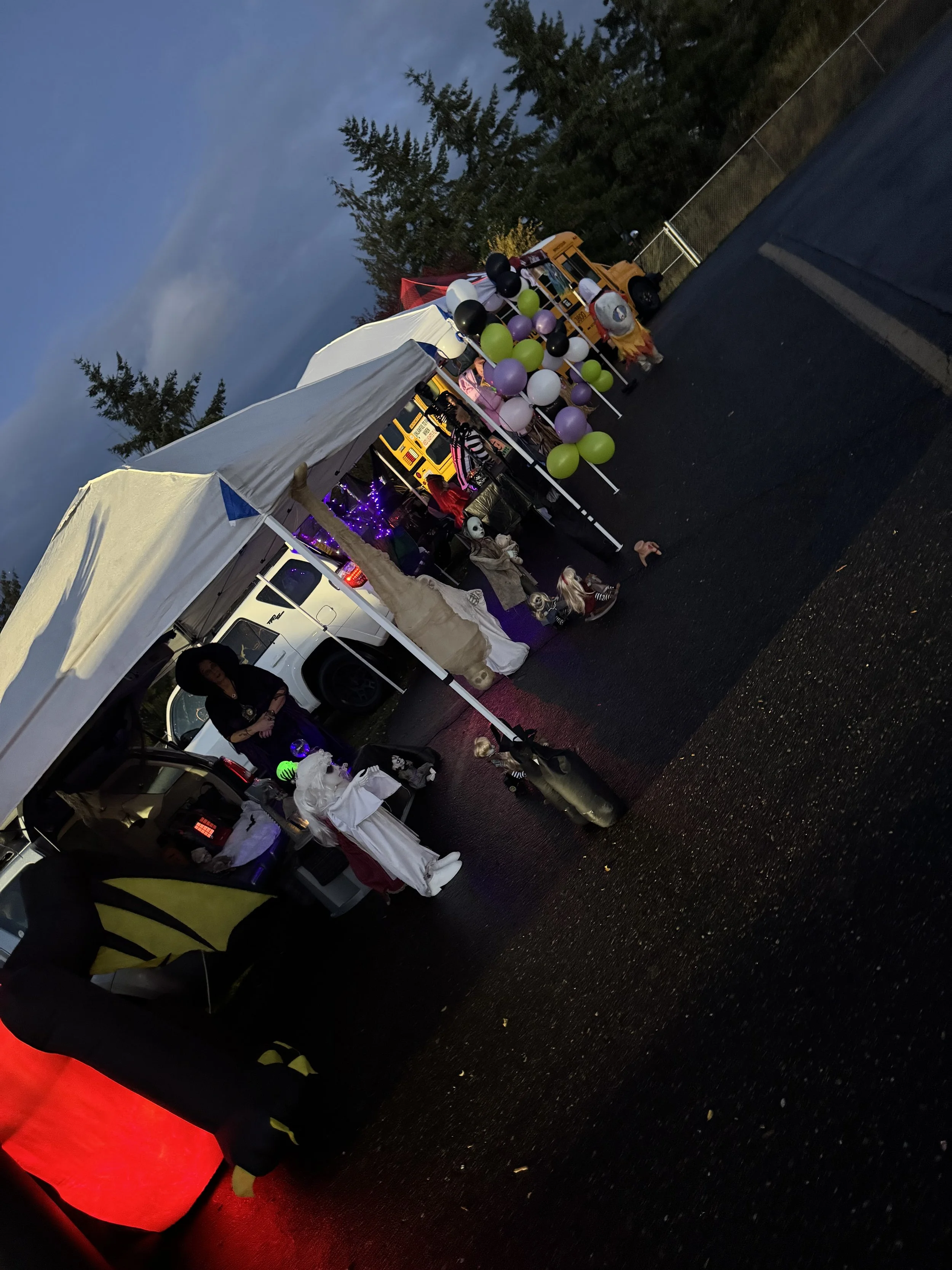 Halloween carnival booth with themed decorations, including balloons, costumes, and a small vehicle with purple lights, set outdoors during dusk.