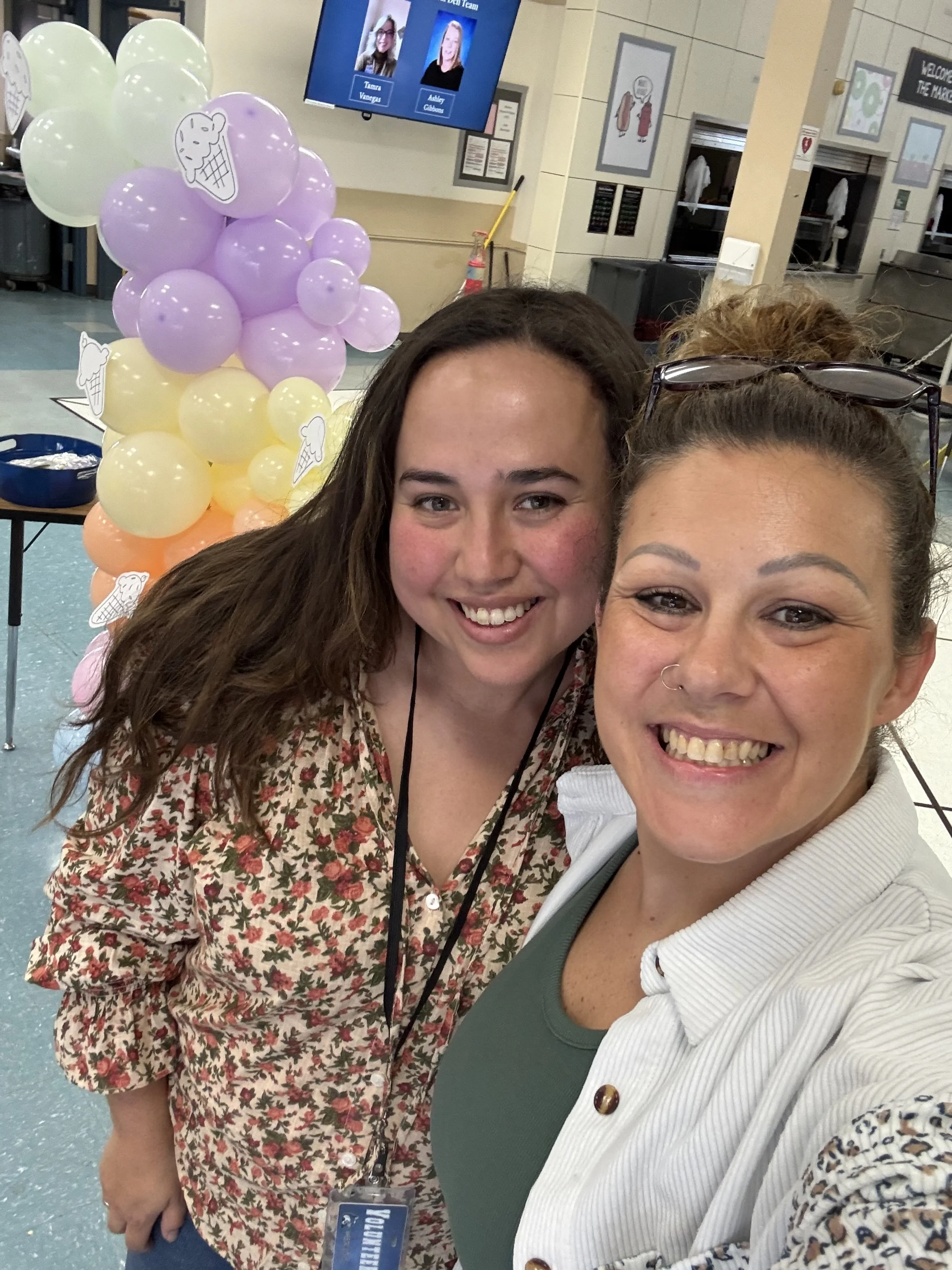 Two smiling women taking a selfie at a colorful ice cream-themed event with balloons and a digital screen in the background.