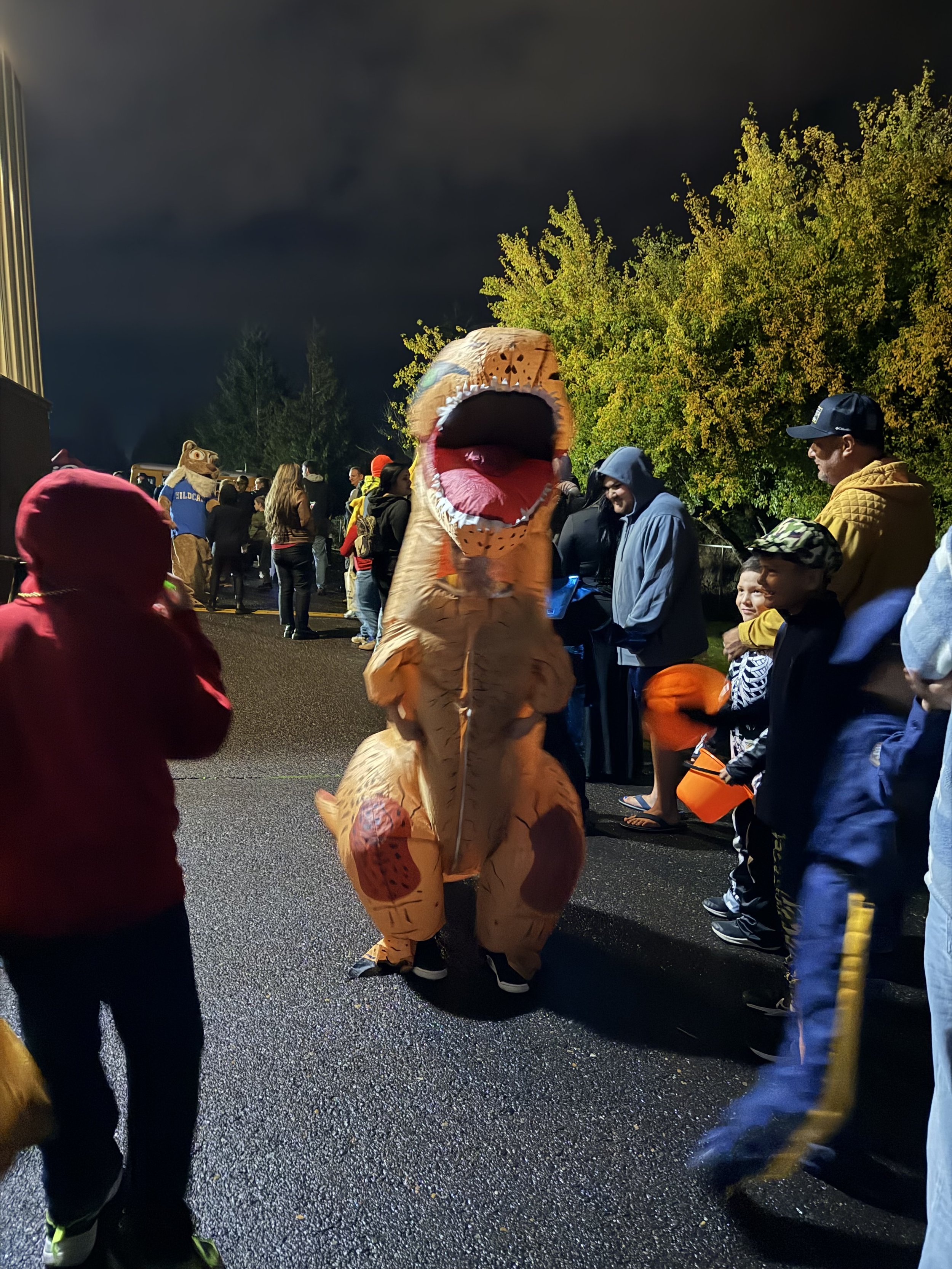 People gather at night outdoors, with a person in a T-Rex costume in the center, and children holding buckets for Halloween candy.