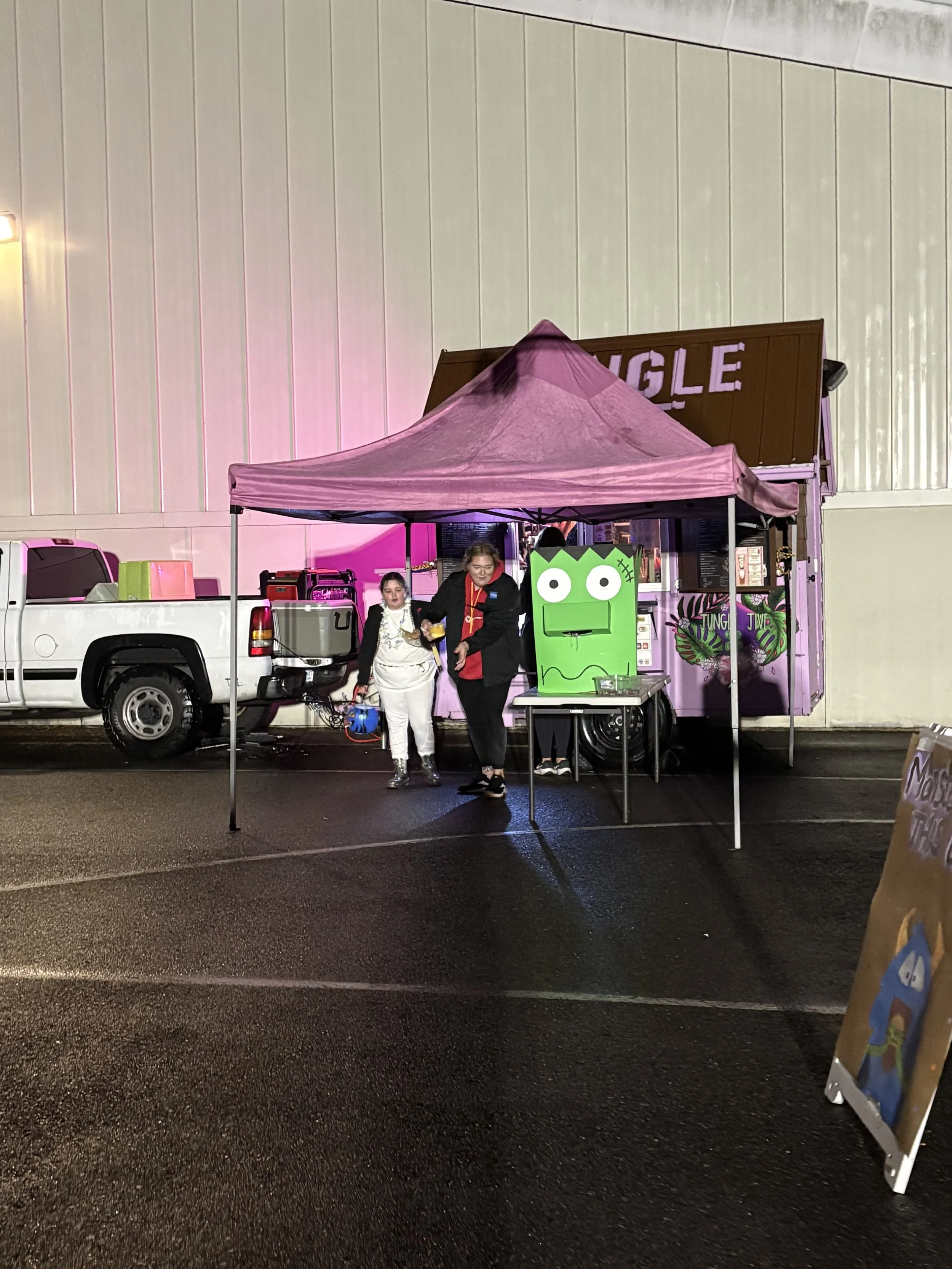 Nighttime outdoor scene of a pink vendor tent with a large green character cutout in front, two people standing under the tent, past a white truck, on a wet pavement, with a large building in the background.