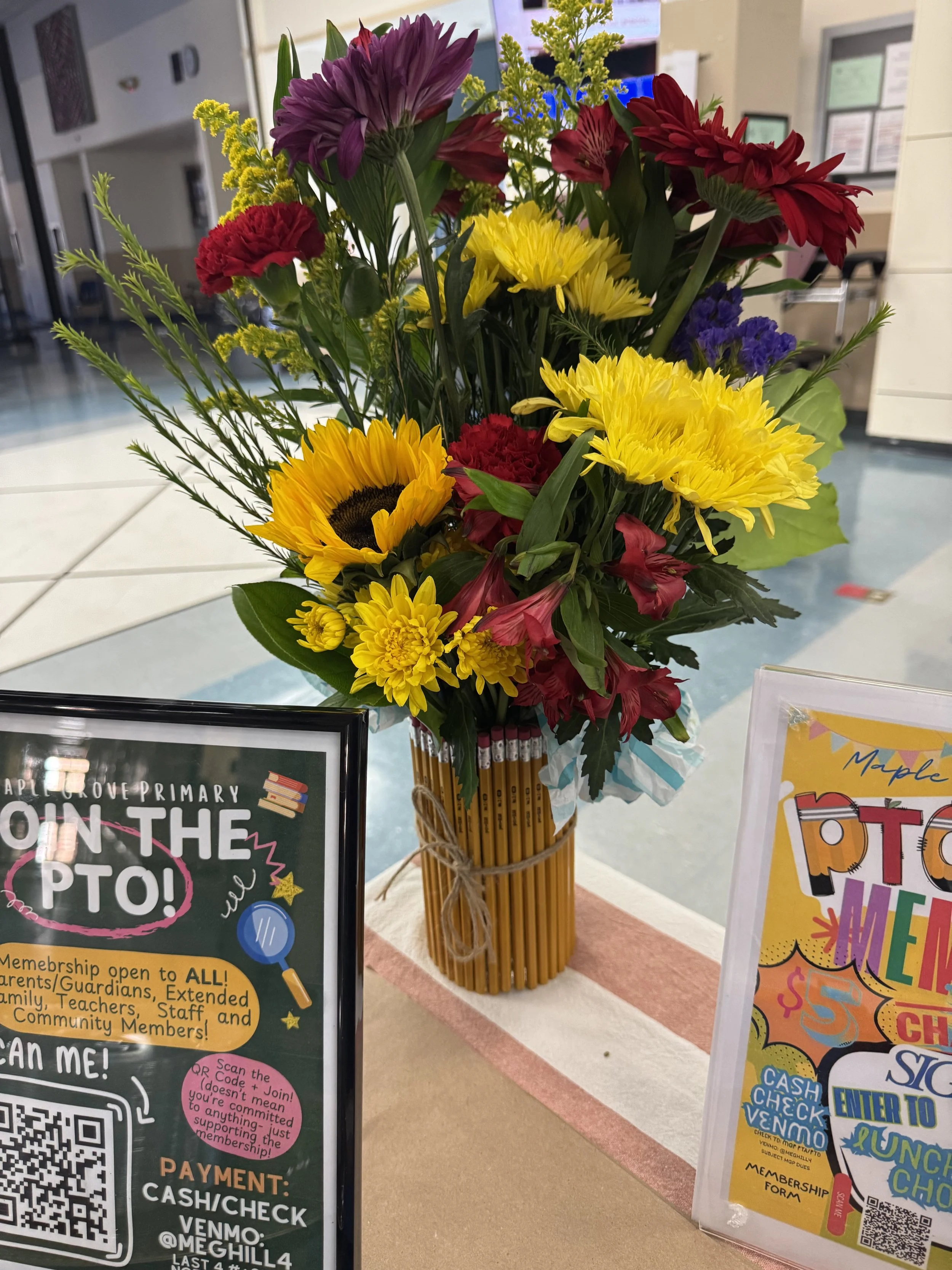 A colorful bouquet of flowers including sunflowers, chrysanthemums, carnations, lilies, and other blooms arranged in a yellow vase with twine, placed on a table with signs for a community event.