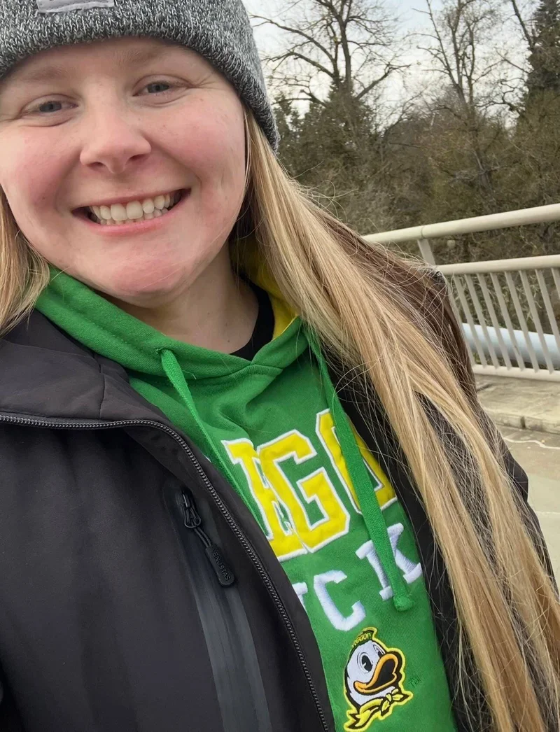 A woman smiling outdoors, wearing a gray knit hat, a green hoodie with yellow and white lettering, and a black jacket. There are trees and a bridge in the background.